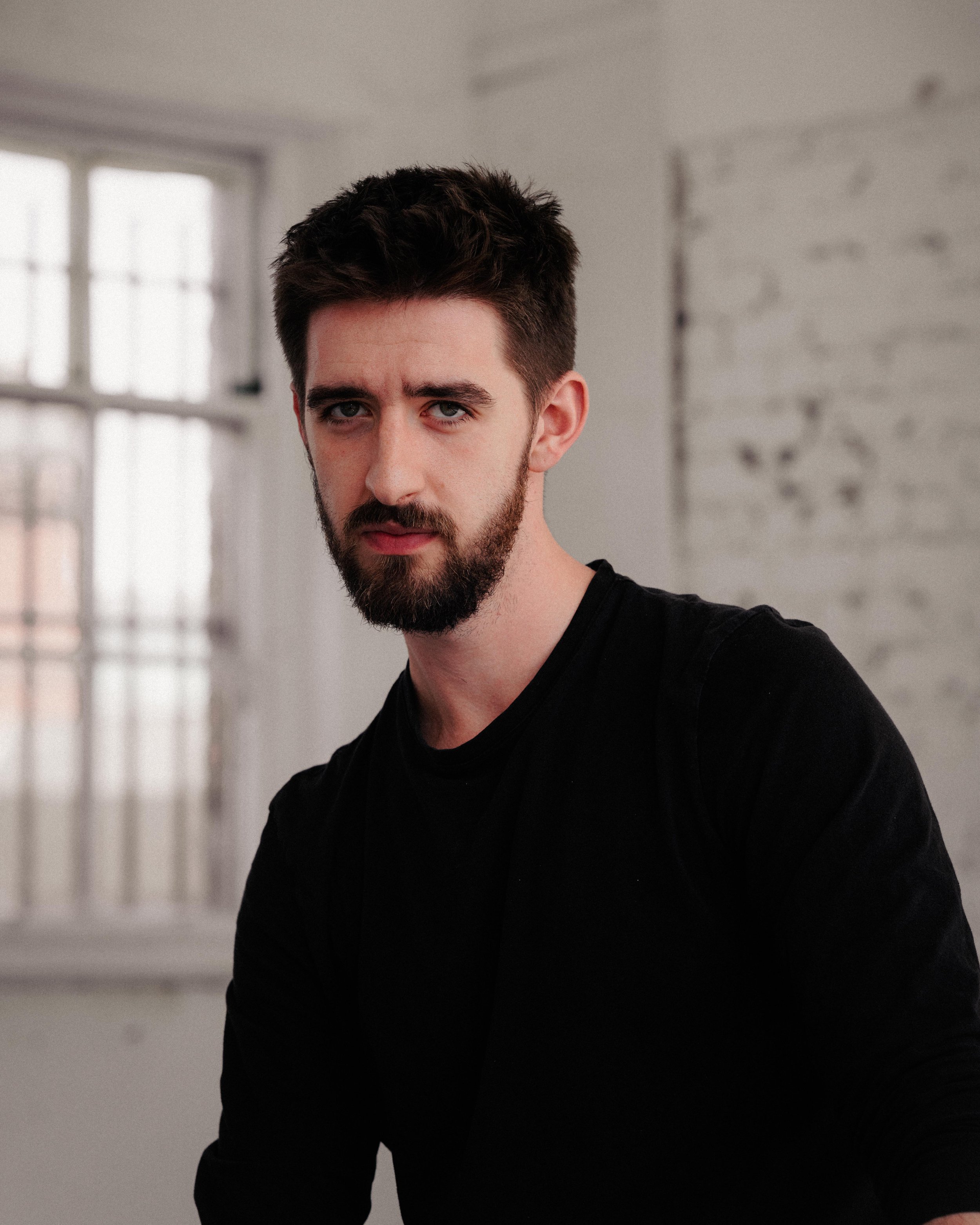 A young man with dark, styled hair, a beard, and blue eyes looking directly at the camera. He is wearing a black shirt. The background is an indoor space with white brick walls and large windows.