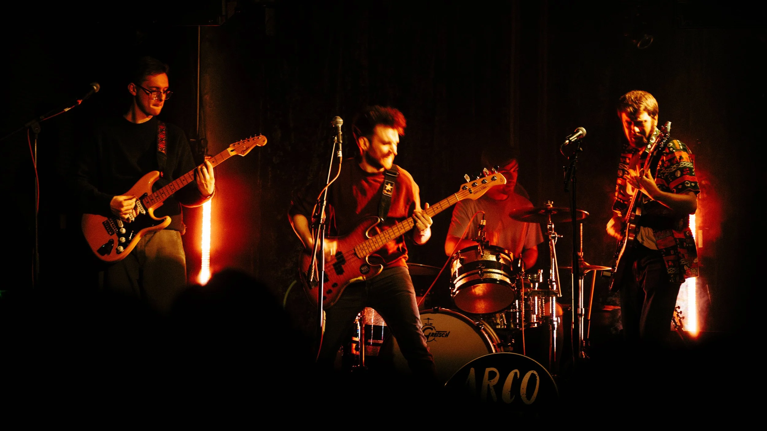 A four-piece band performing live on stage with guitars, bass, and drums, under warm red and orange stage lights.