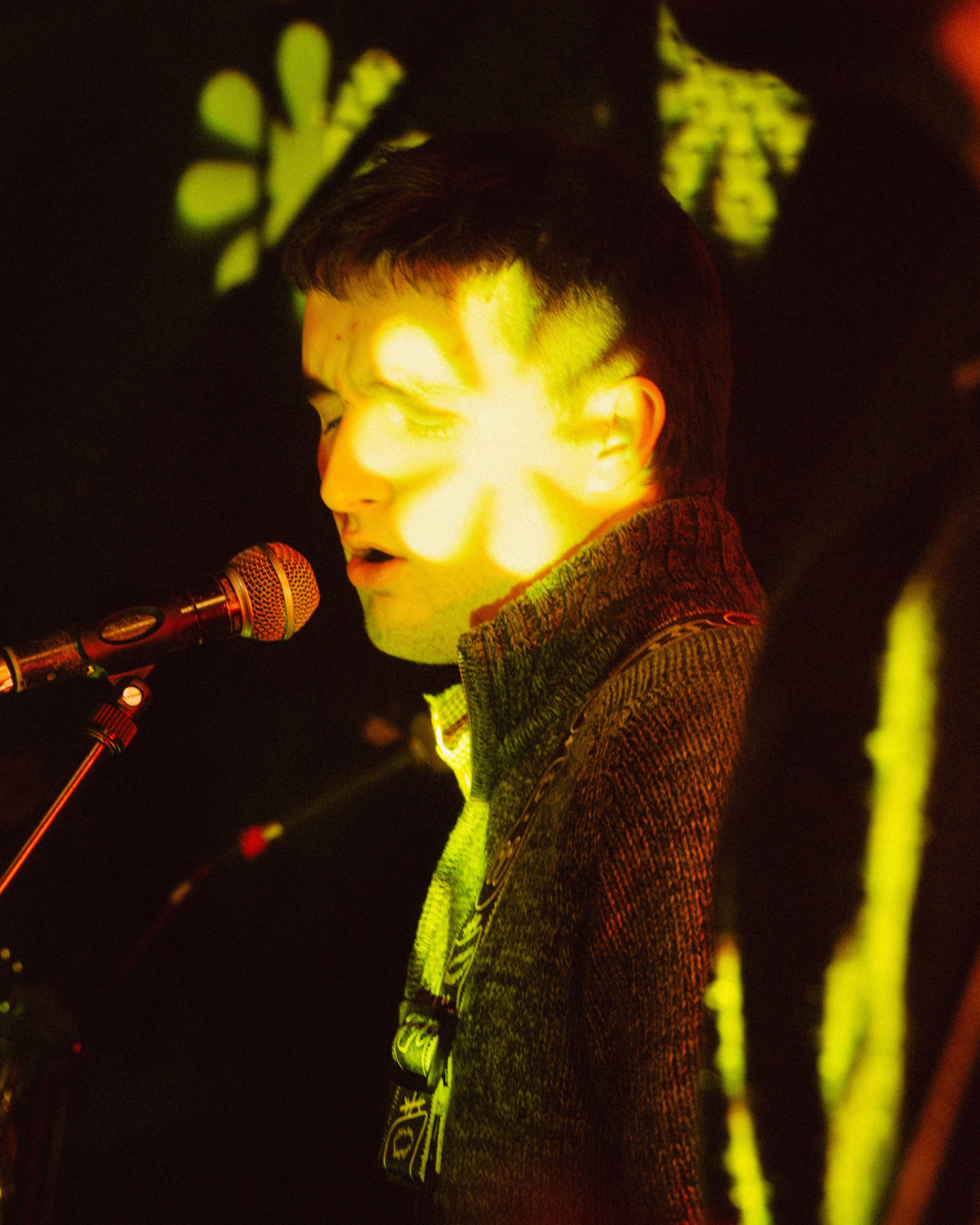 A young man with dark hair and closed eyes singing into a microphone under yellow and green stage lights.