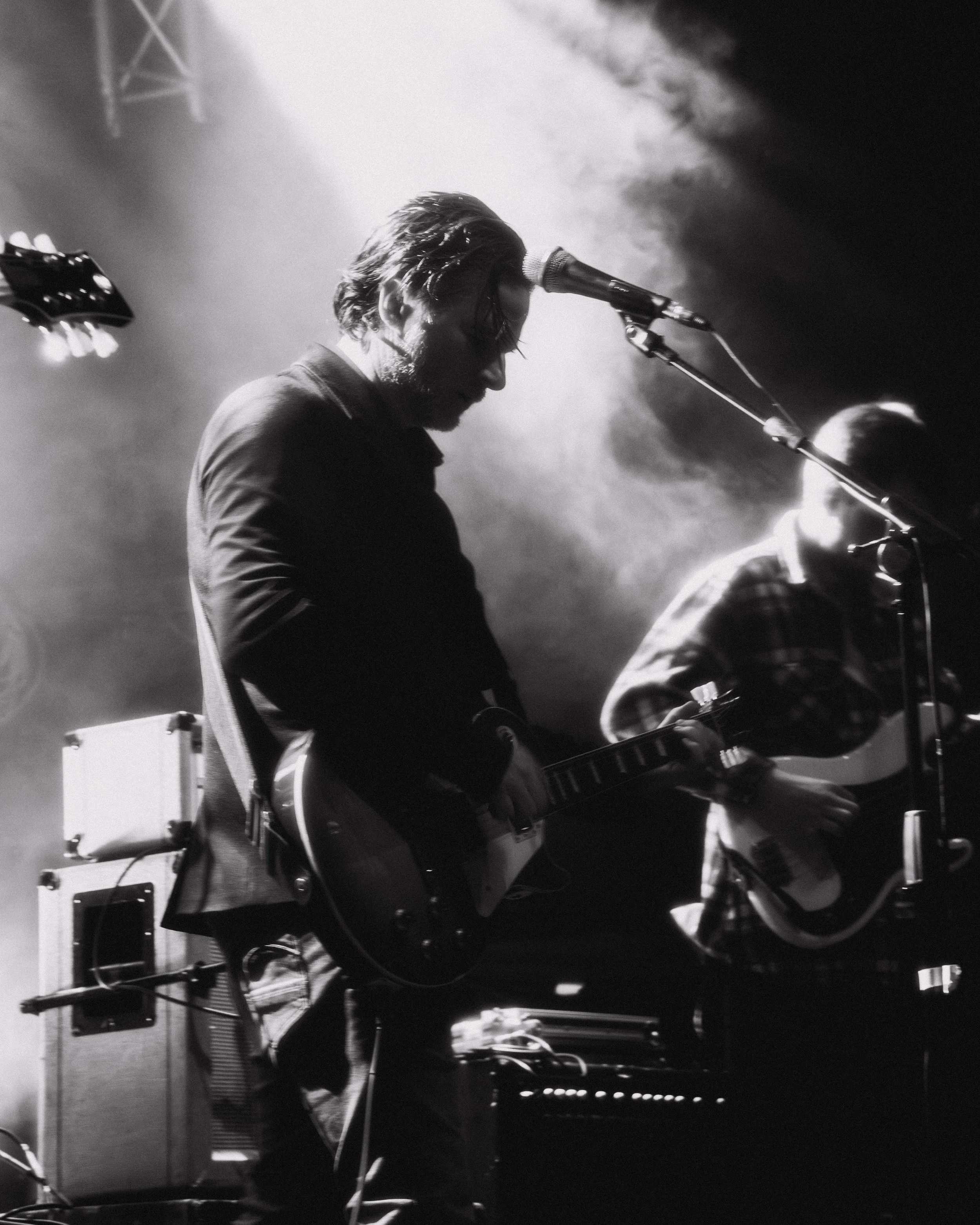 Musicians performing on stage, playing guitars, in black and white, with stage lighting and smoke effects.
