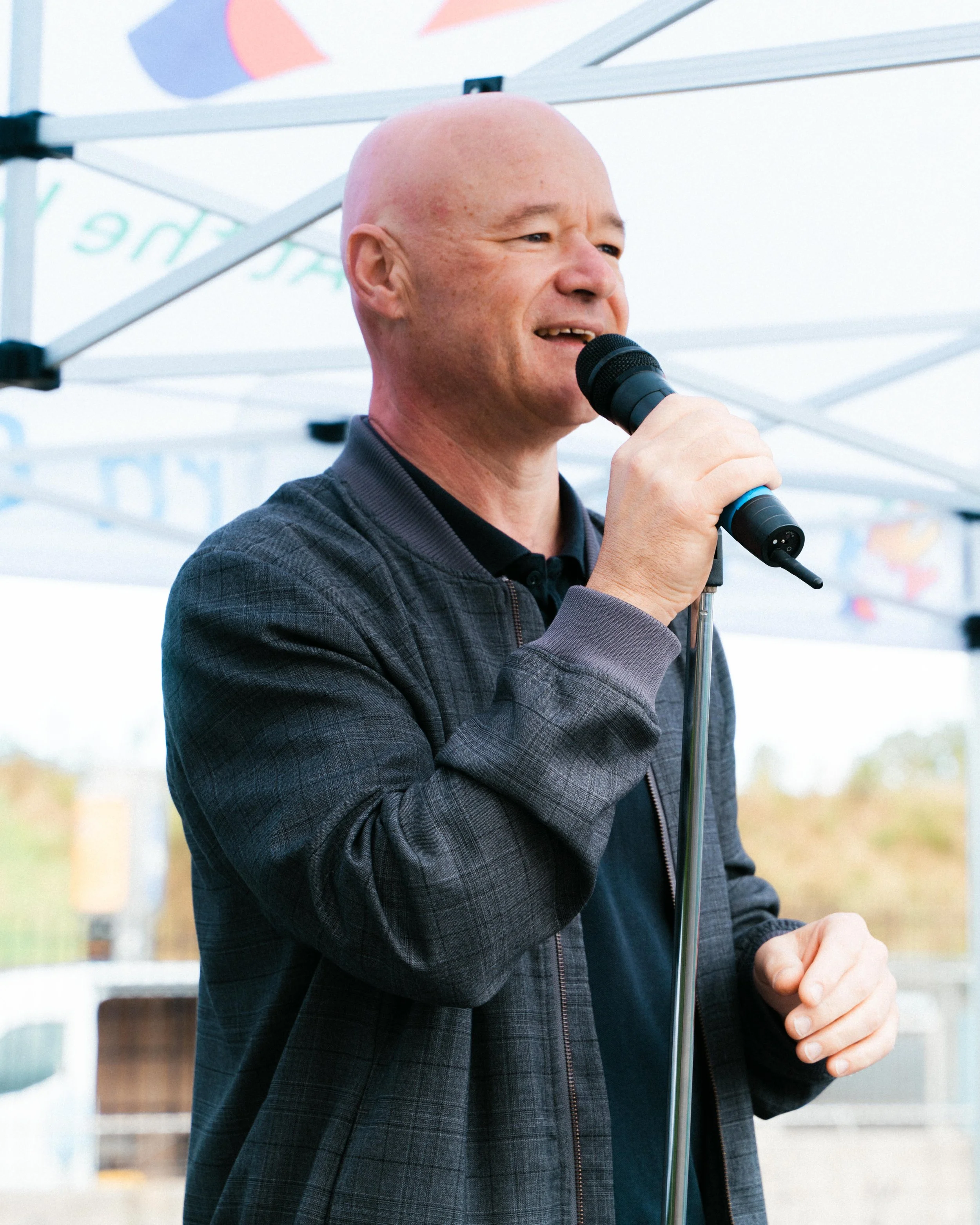 A man with a bald head is holding a microphone and appears to be speaking at an outdoor event.