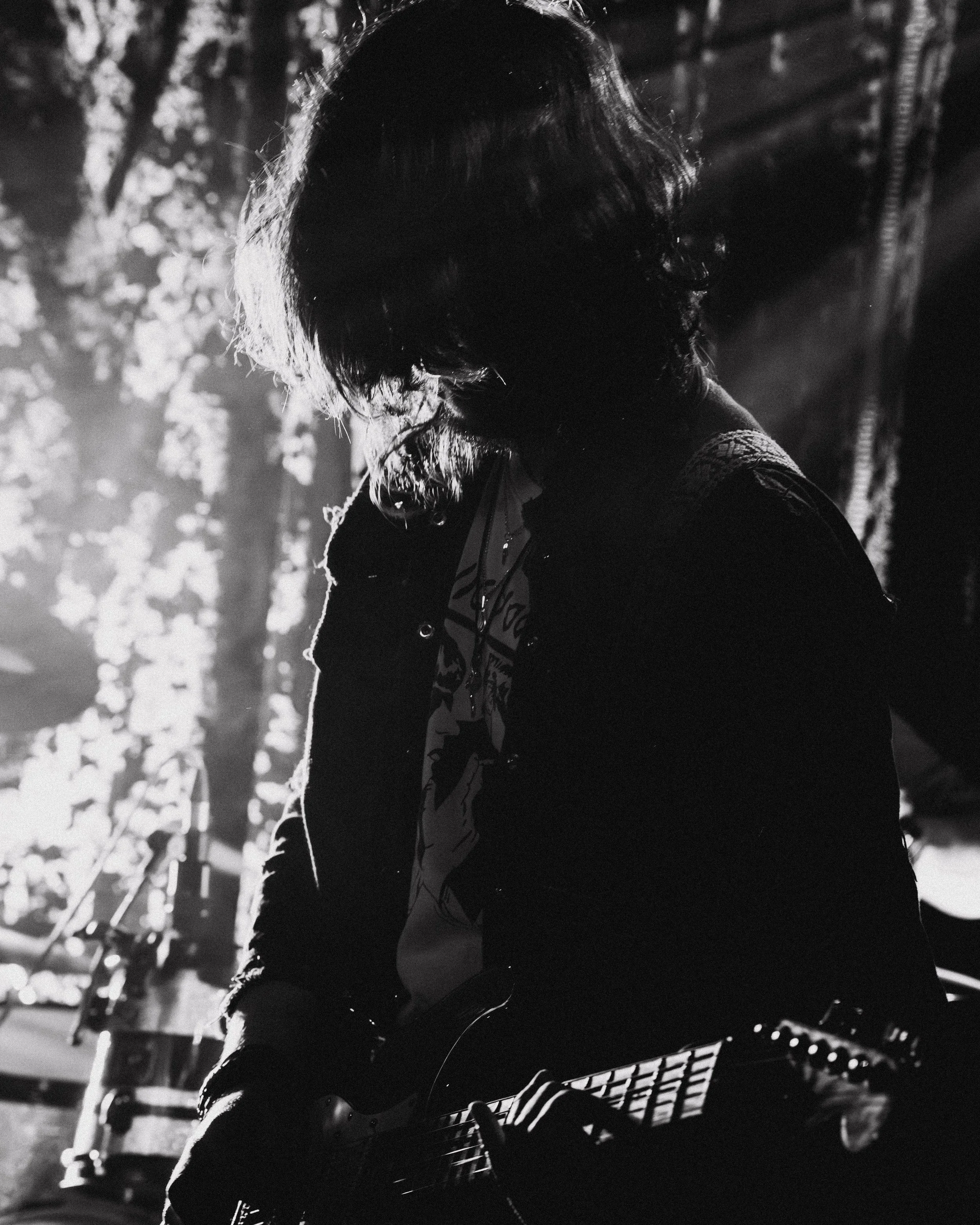 A person with shoulder-length hair playing an electric guitar on stage, backlit by bright lights, in black and white.