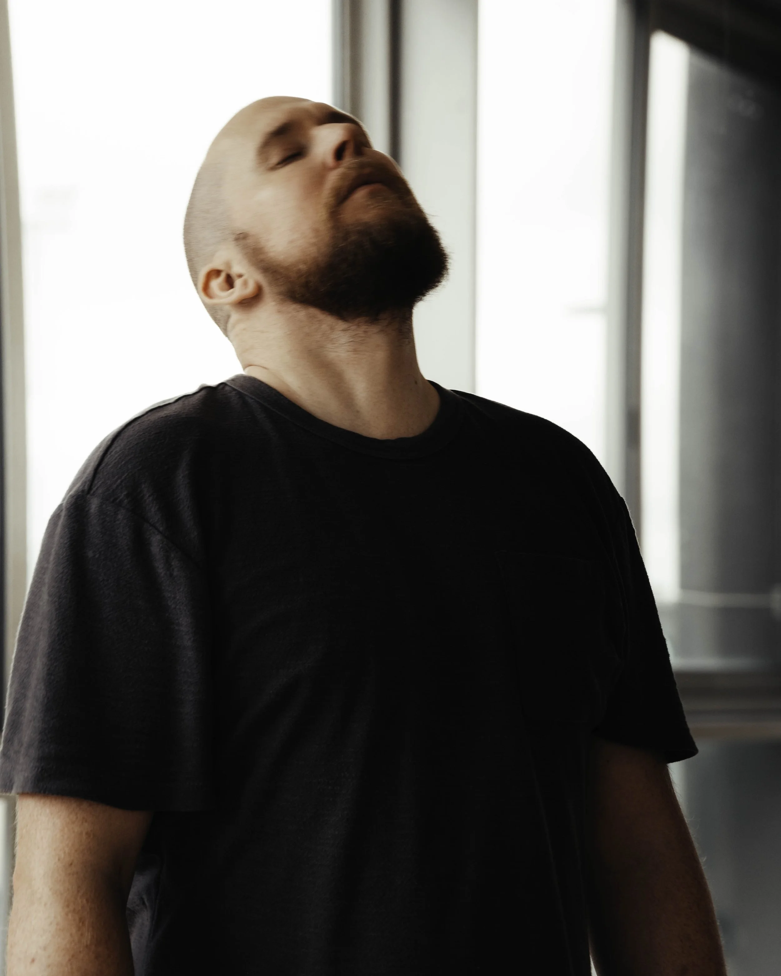 Close-up of a man with a shaved head and beard, wearing a black shirt, standing indoors near a window with light coming through.