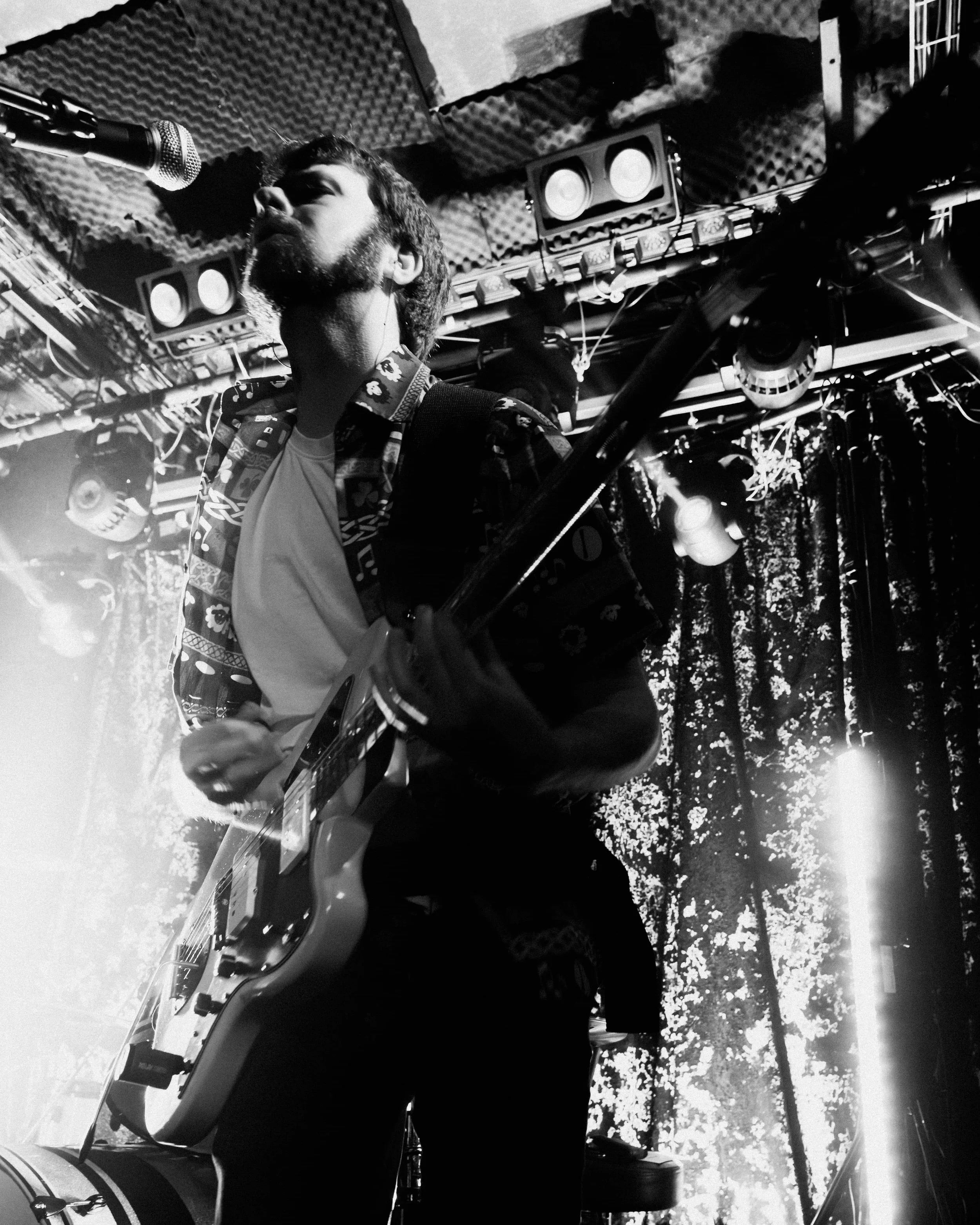 A black and white photo of a male musician playing an electric guitar on stage, near a microphone, with stage lights and sound equipment overhead.