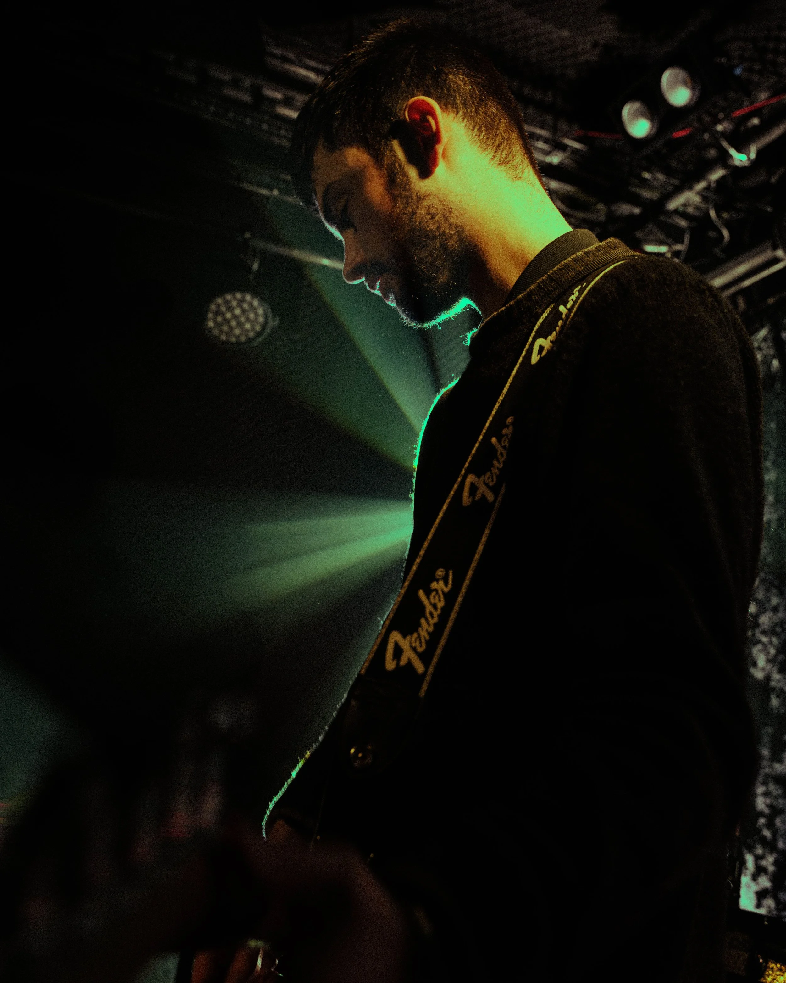 A young man with dark hair and beard playing a guitar on stage, illuminated by green stage lights in a dark setting.