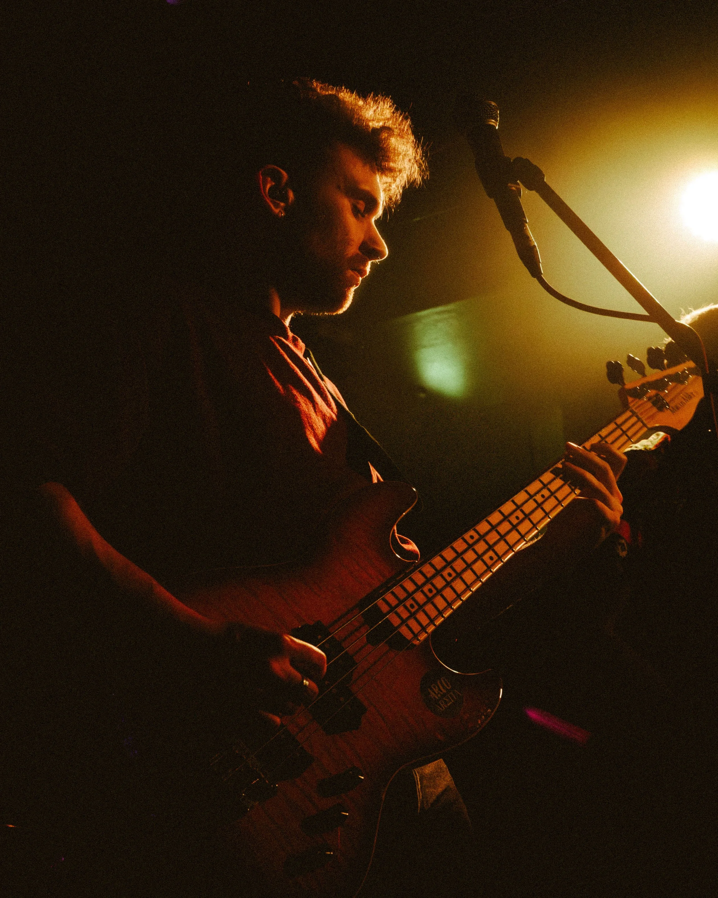 A musician playing an electric bass guitar during a live performance, illuminated by stage lighting.