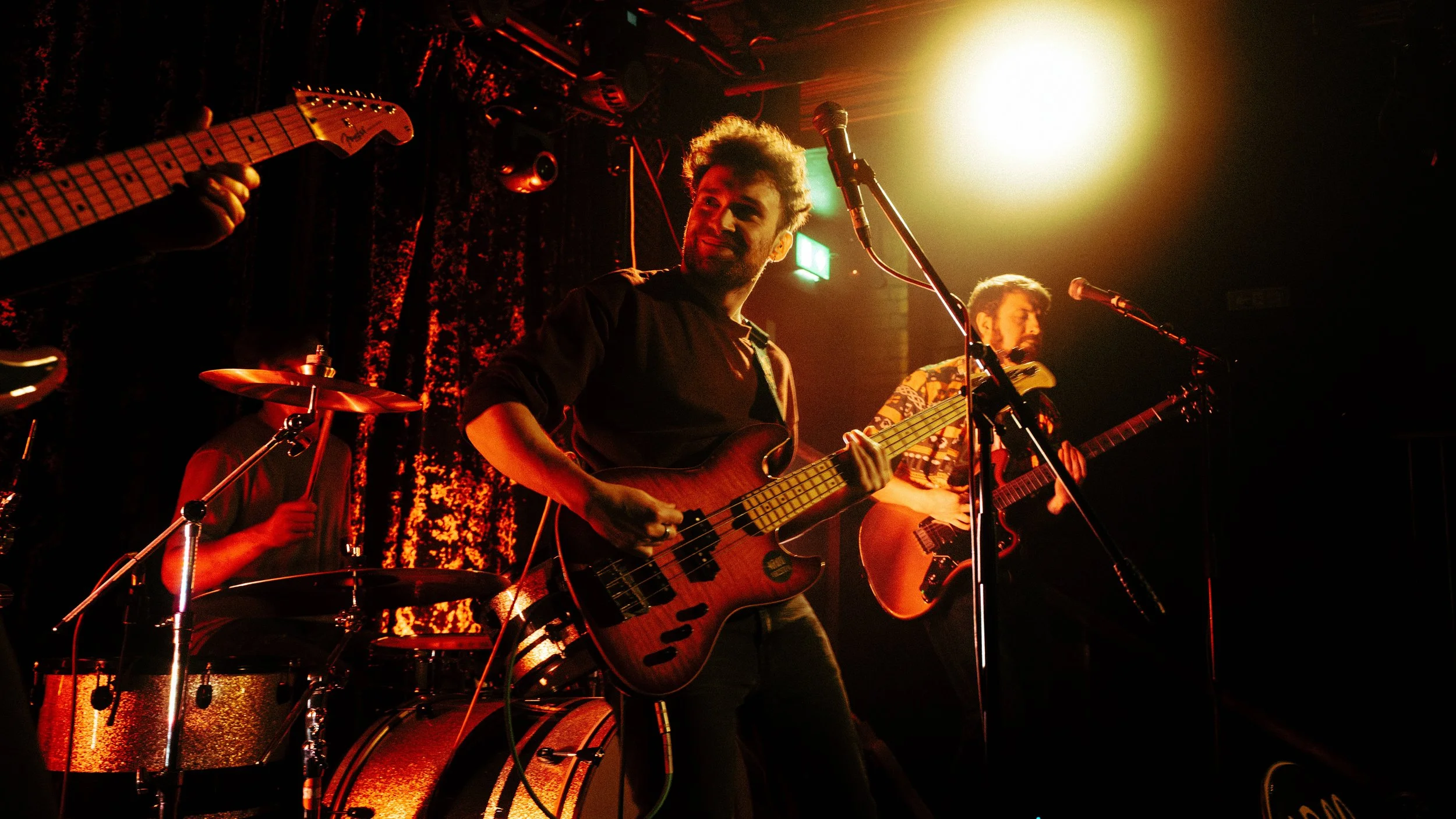 Musicians performing on stage, with one playing bass guitar, another playing an acoustic guitar, and a drummer in the background, illuminated by stage lights.