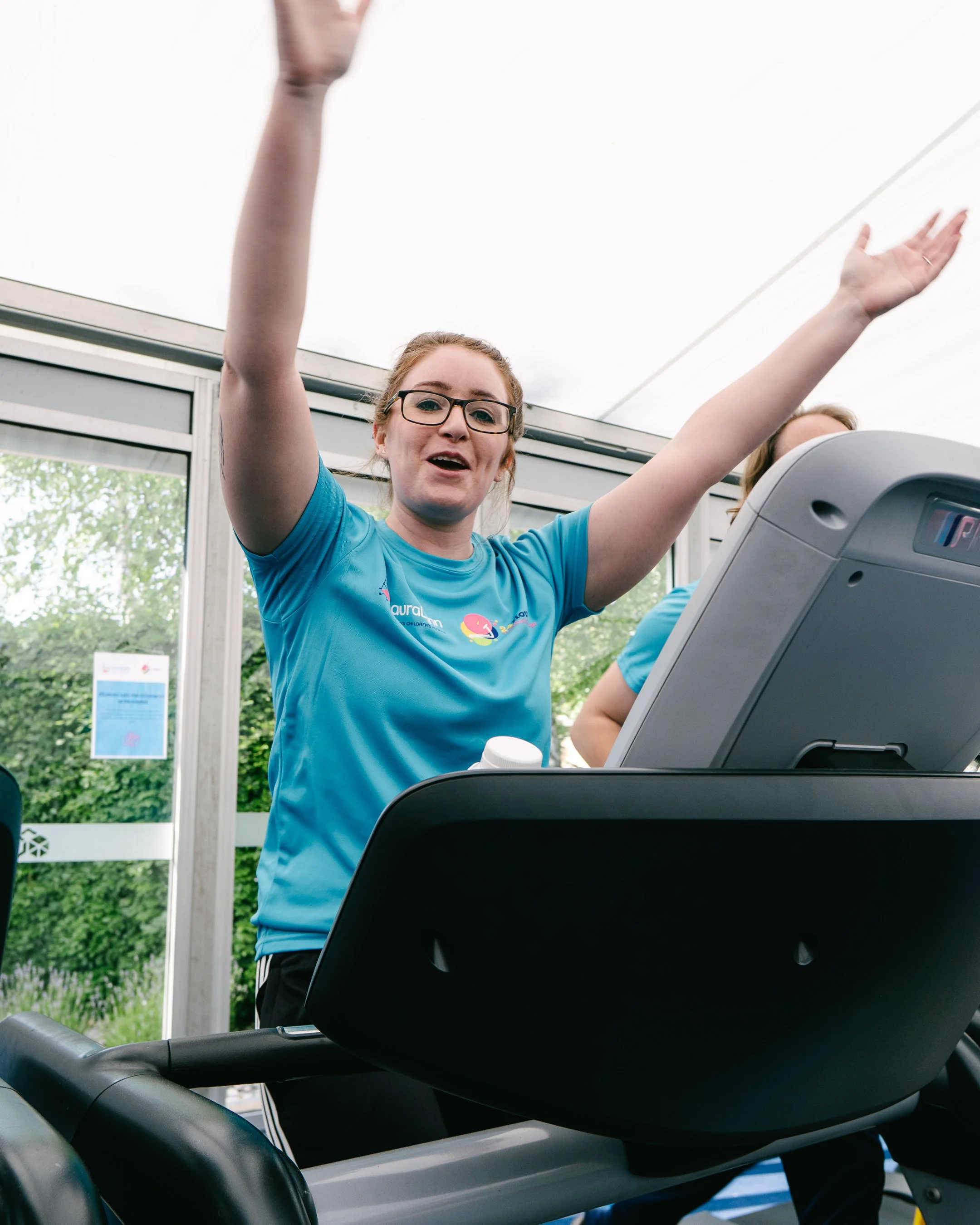A young woman with glasses and a blue shirt is standing on a treadmill, raising her arms in a gym or wellness center, with another person also on a treadmill visible in the background.