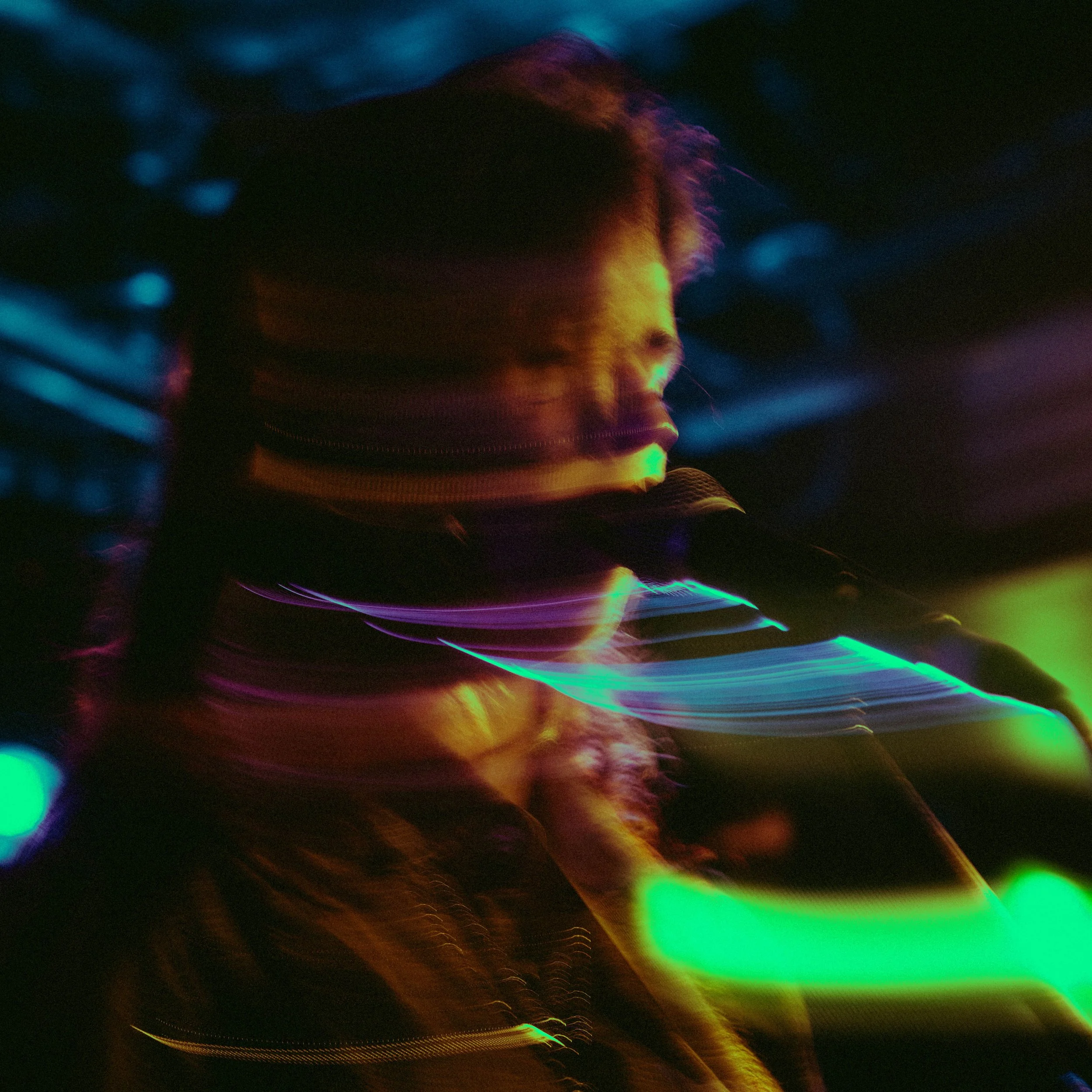 A person is singing or speaking into a microphone on stage, with colorful and blurry lights creating a vibrant, abstract background.