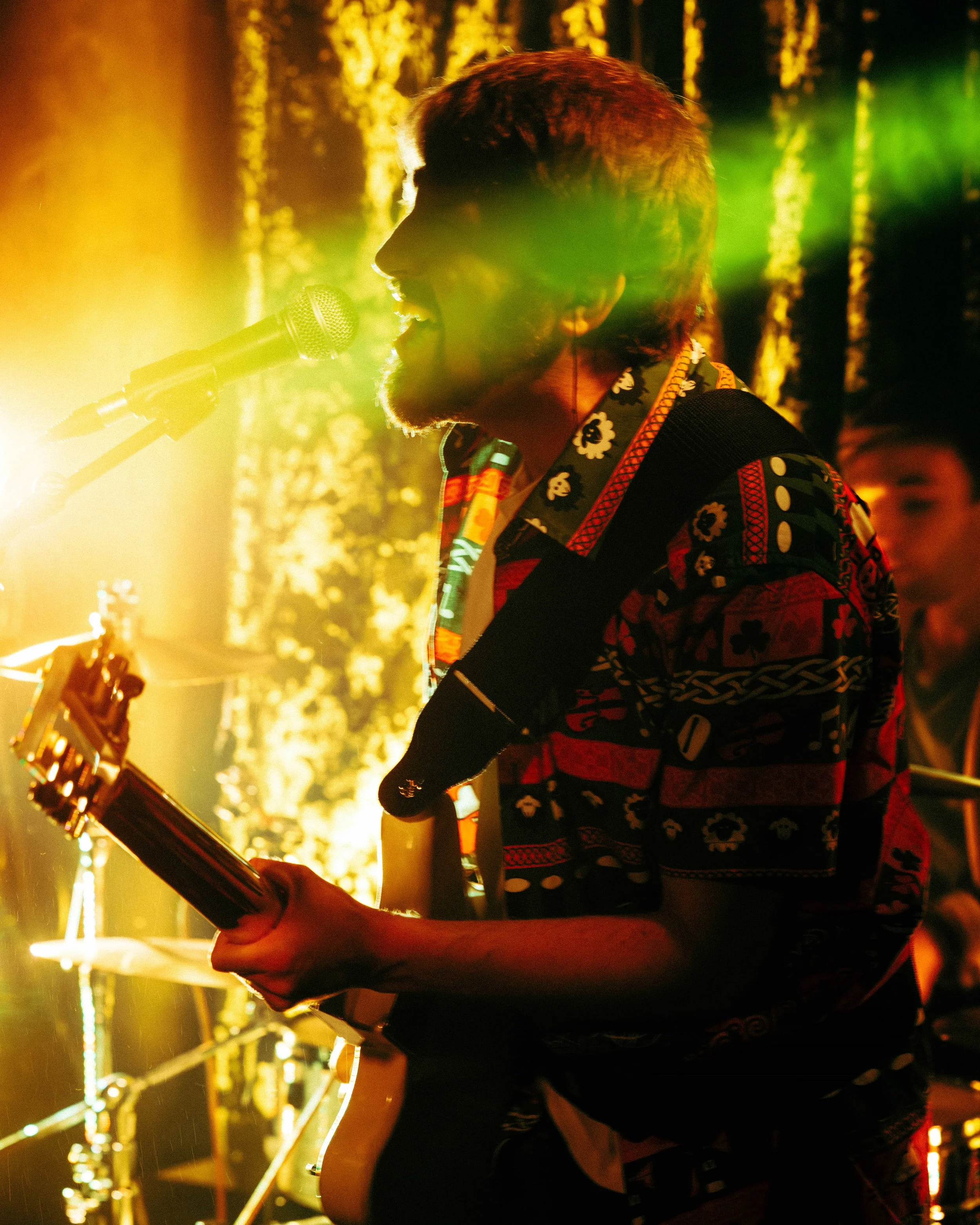 Musician singing into a microphone while playing an electric guitar during a live performance with colorful stage lighting.
