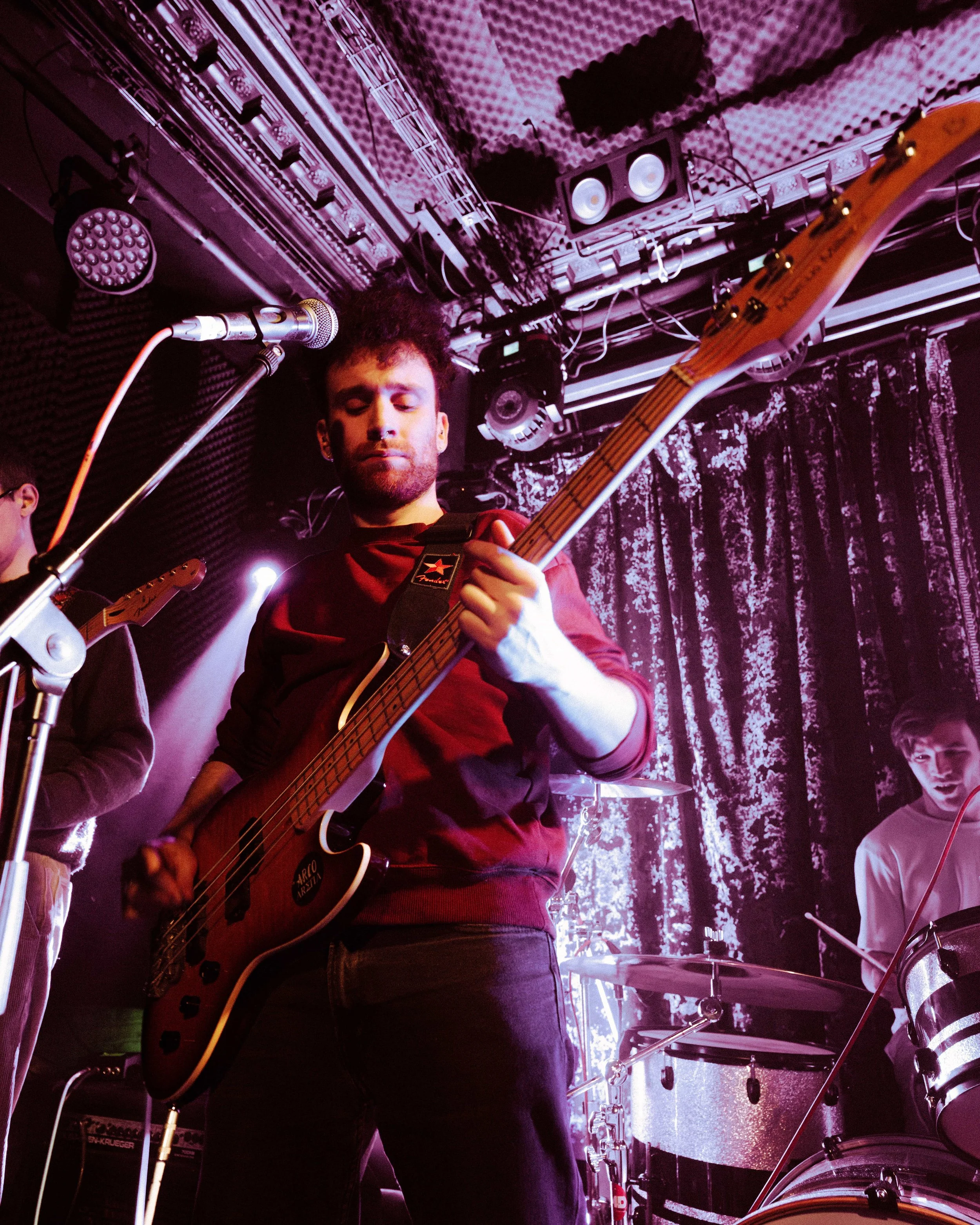 A man playing bass guitar on stage during a live music performance, with a woman in the background playing drums and another person partially visible.