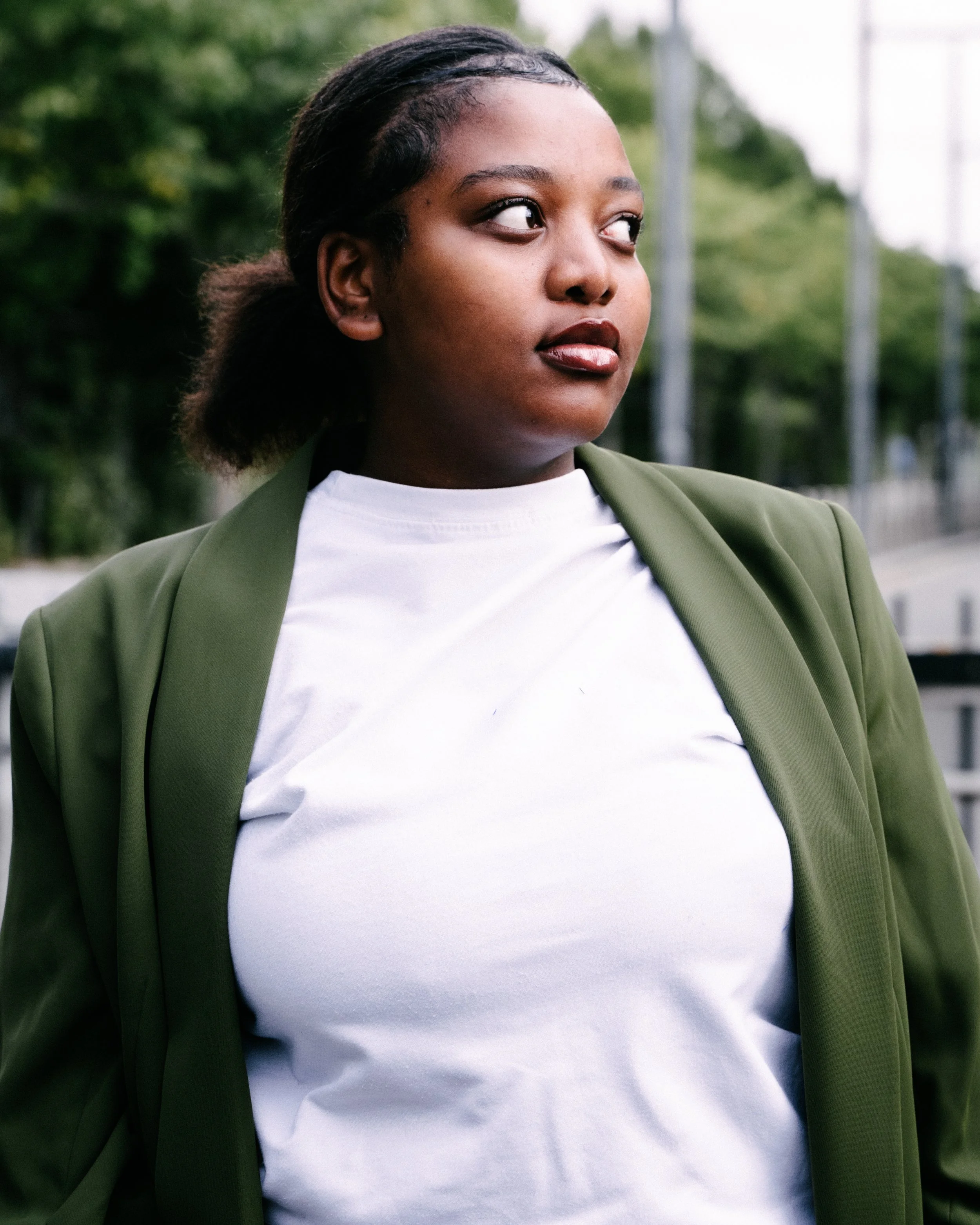 A close-up of a young Black woman wearing a white t-shirt and an olive green blazer, outdoors with trees and a fence in the background.
