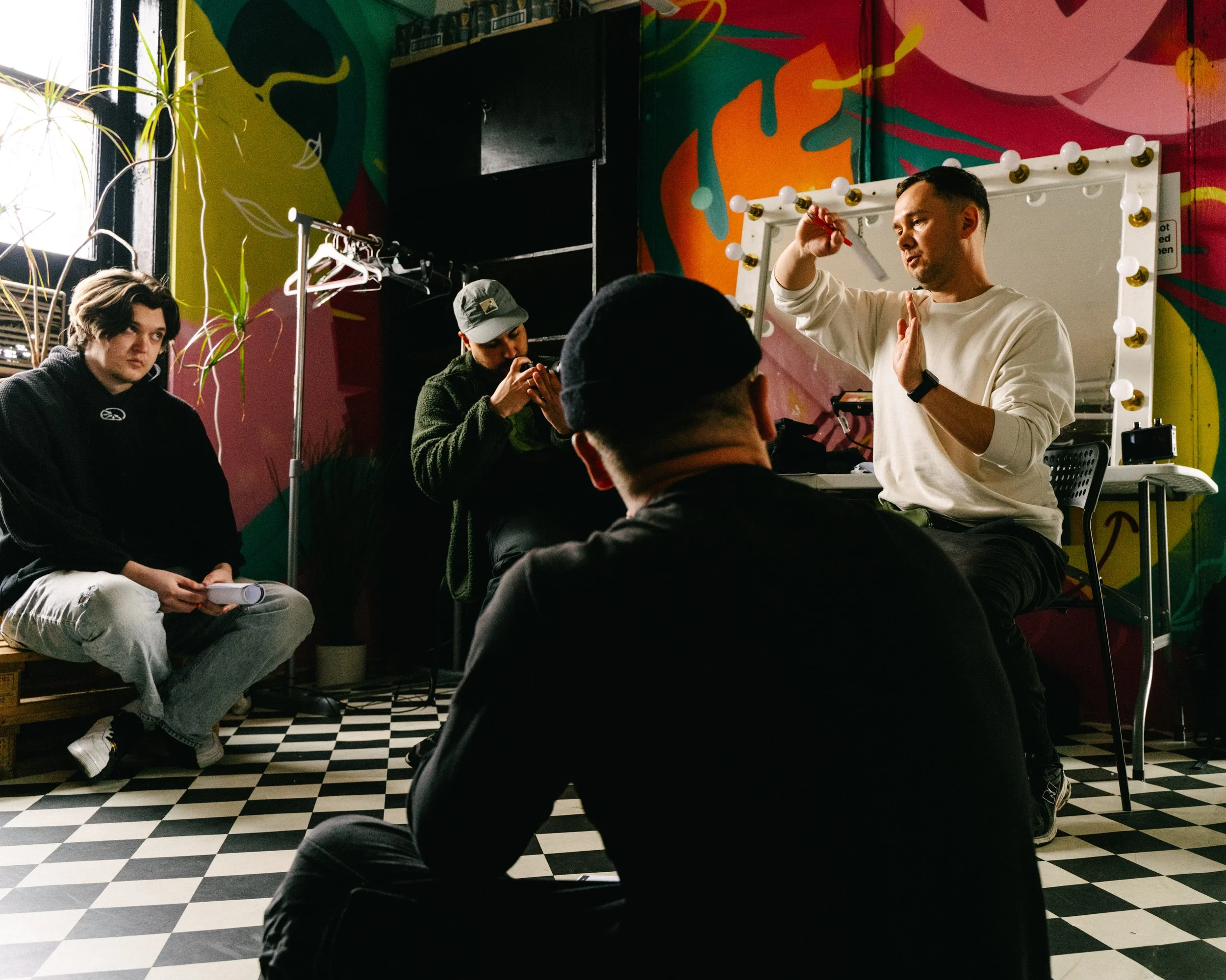 A man is standing in front of a mirror with light bulbs around the frame, applying makeup or grooming. Several people are seated in the foreground, with one person working on a phone. The background features a colorful wall with abstract patterns and