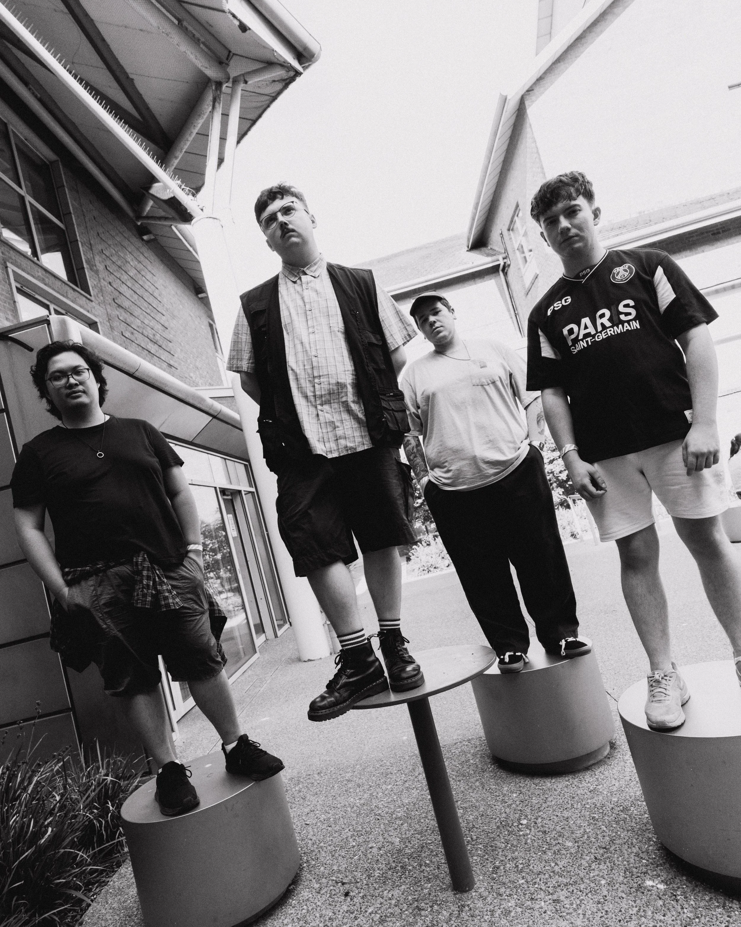 Four young men standing behind large planters in front of a building, wearing casual clothing including T-shirts, shorts, and sneakers, with one wearing glasses and another wearing a cap.