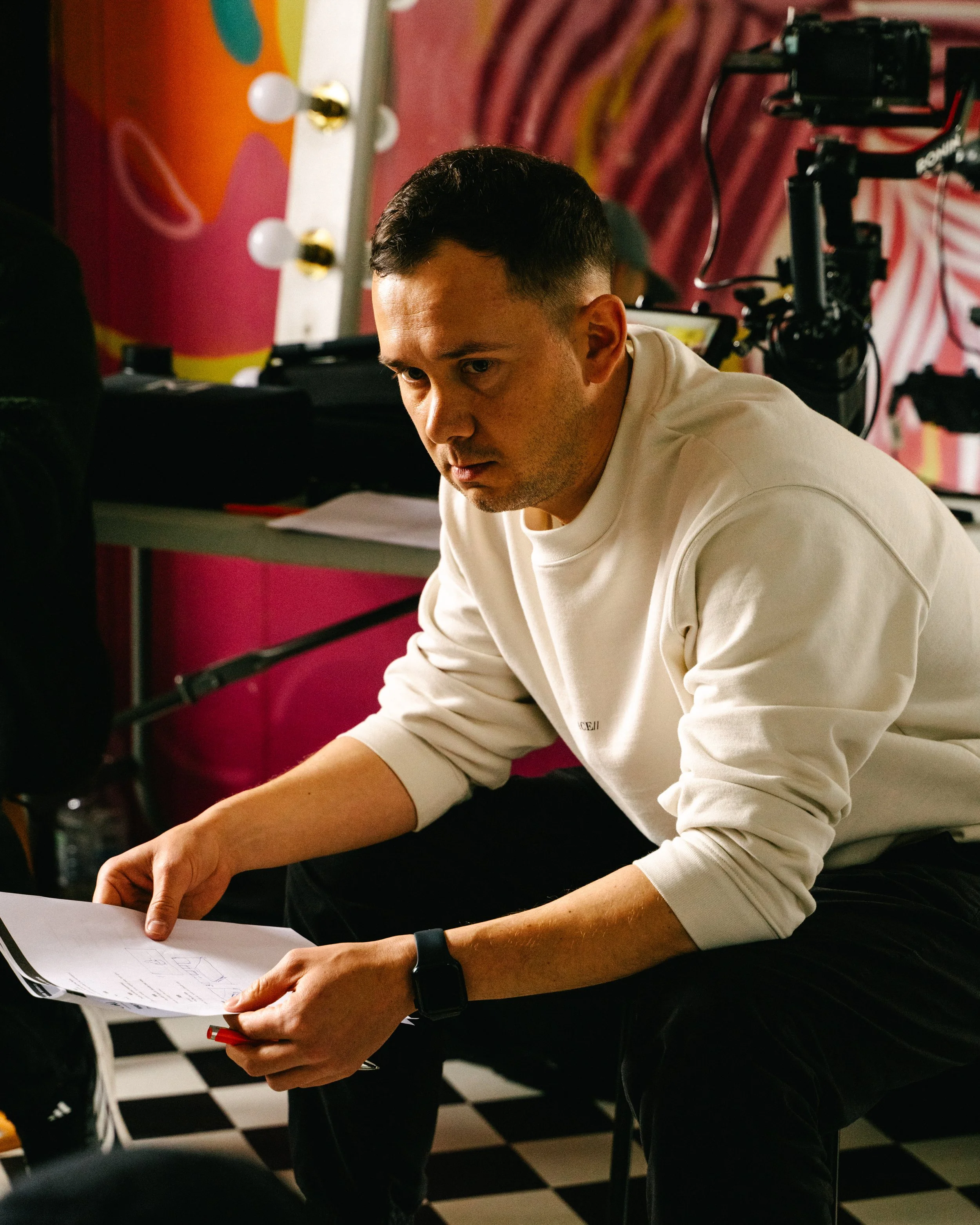 A man with short black hair, wearing a white long-sleeve shirt and a smartwatch, sitting at a table with a document in his hand, appears to be working or reviewing something. The background features a colorful polka dot and striped pattern on the wal