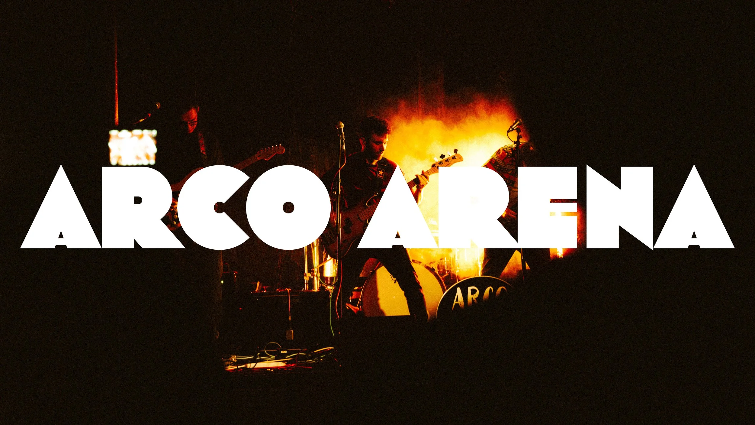 A band performing on stage with the word 'ARCO ARENA' in bold white letters overlaying the scene. The stage is dark with a fiery background illuminating the band members.