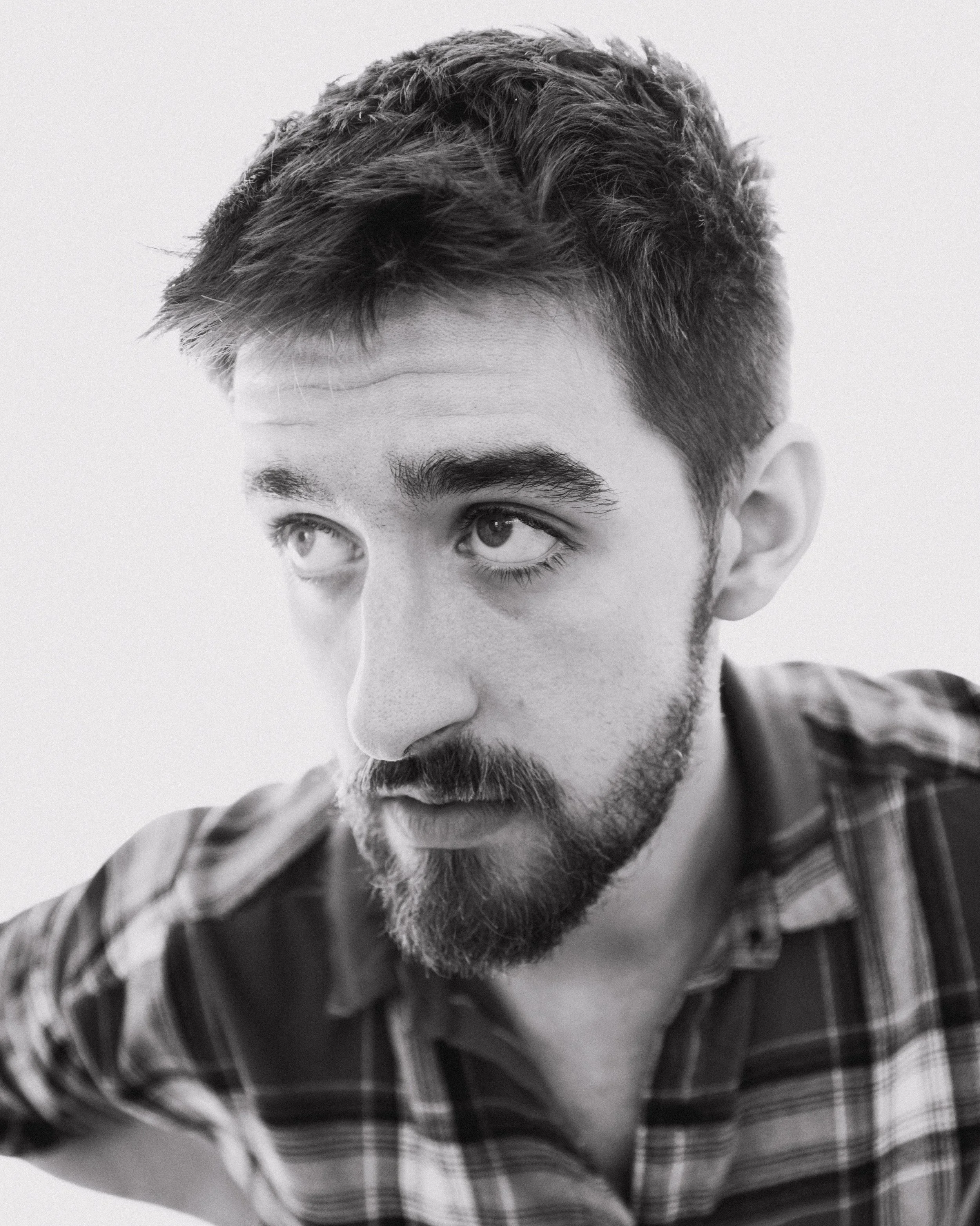 A black and white close-up photo of a young man with a beard and short, wavy hair, looking downwards, wearing a plaid shirt.