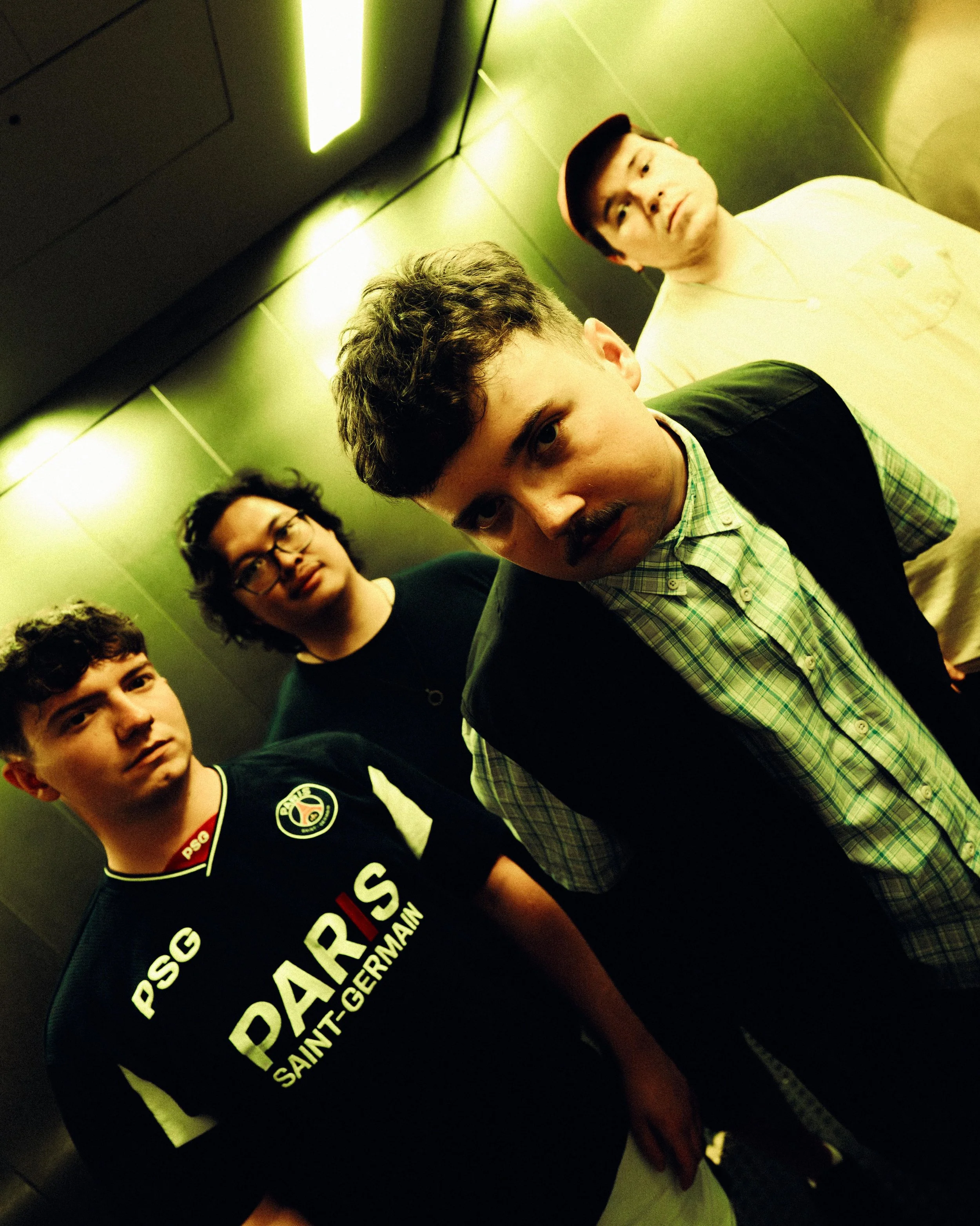 Four young men standing in an elevator, three facing forward and one in front, smiling and looking at the camera. The elevator has greenish walls and bright overhead lighting.