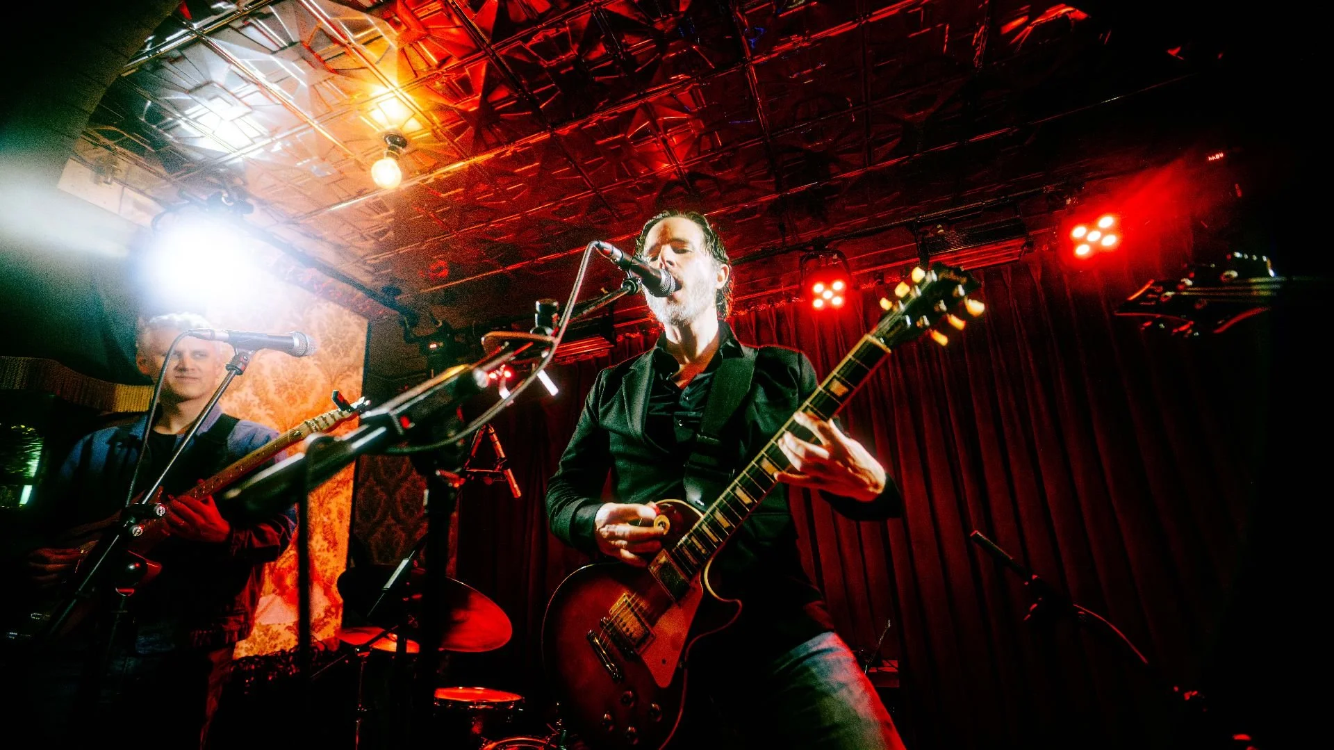 Musicians performing live on stage, with one playing an electric guitar and singing into a microphone, and another playing a guitar in front of a dark red curtain with stage lights overhead.