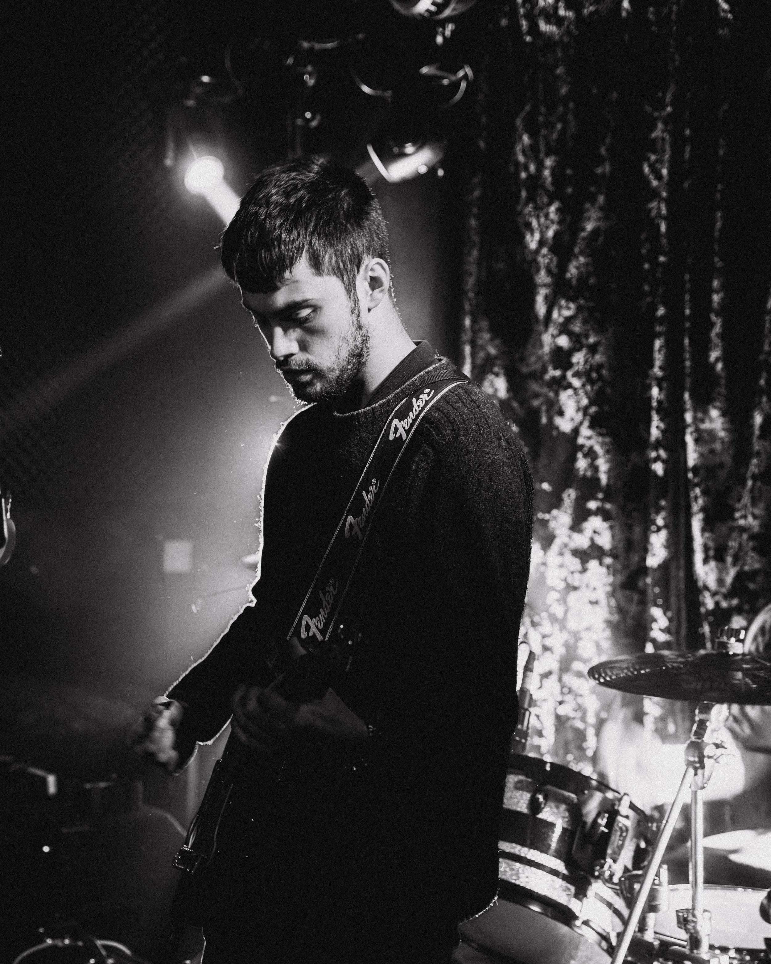 A young man playing an electric guitar on stage, with a drum set visible in the background, under stage lighting.
