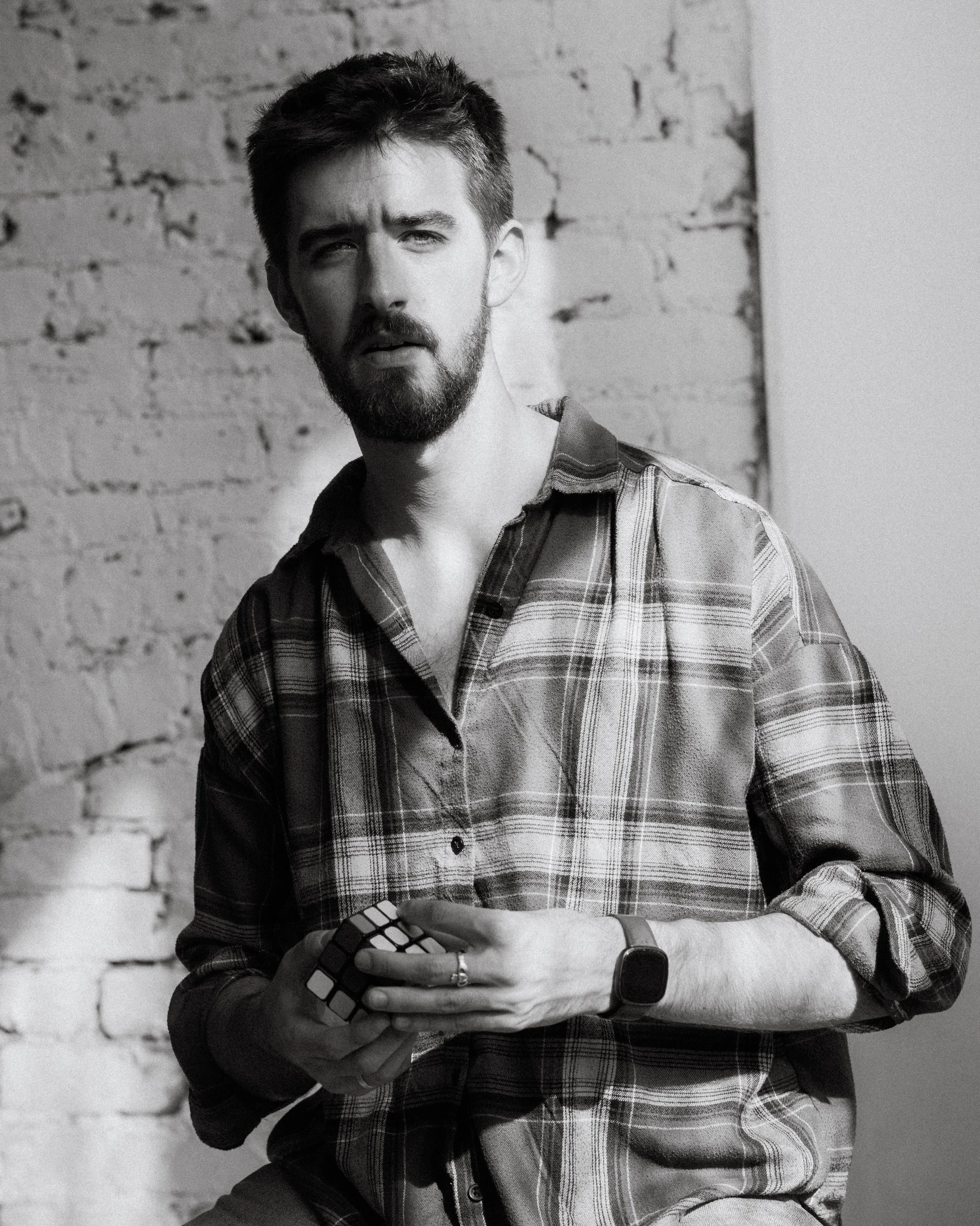 A young man with a beard and short hair wearing a plaid shirt, holding a Rubik's cube, standing against a brick wall with a serious expression.