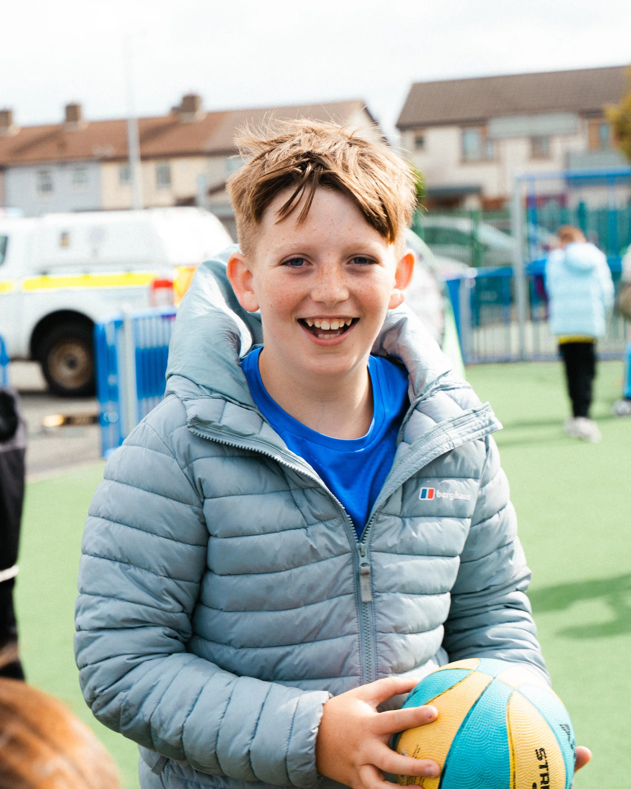 A boy with reddish hair, freckles, and blue eyes smiling while holding a yellow and blue volleyball at an outdoor playground or sports field, with blurred houses and a fence in the background.