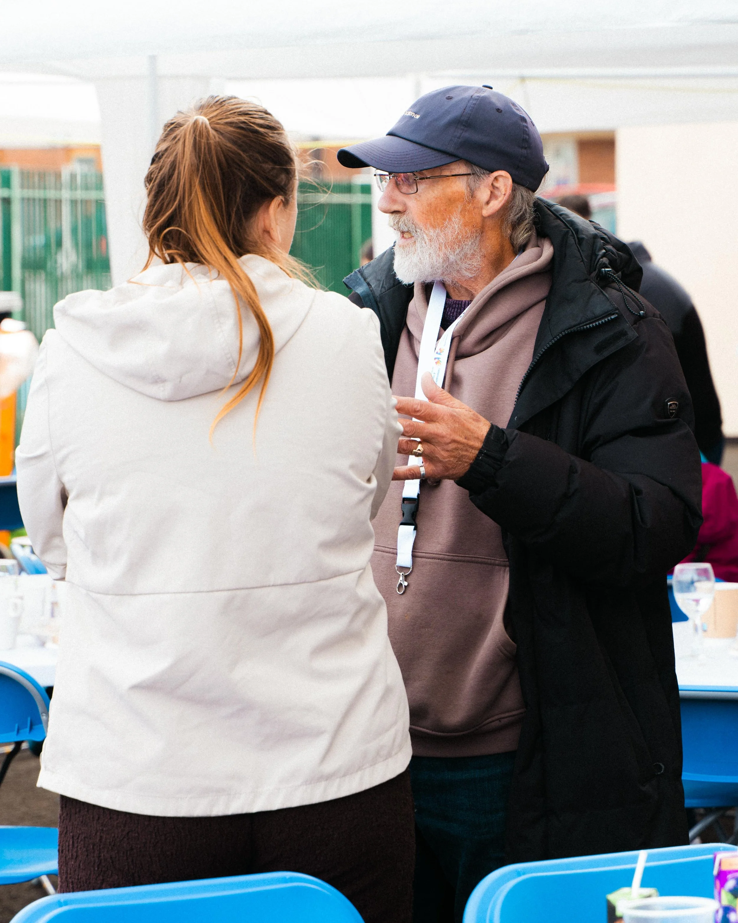 An older man with glasses and a beard, wearing a navy cap, hoodie, and black jacket, talking to a woman with long reddish-brown hair, wearing a white jacket, at an outdoor event with tables and chairs.