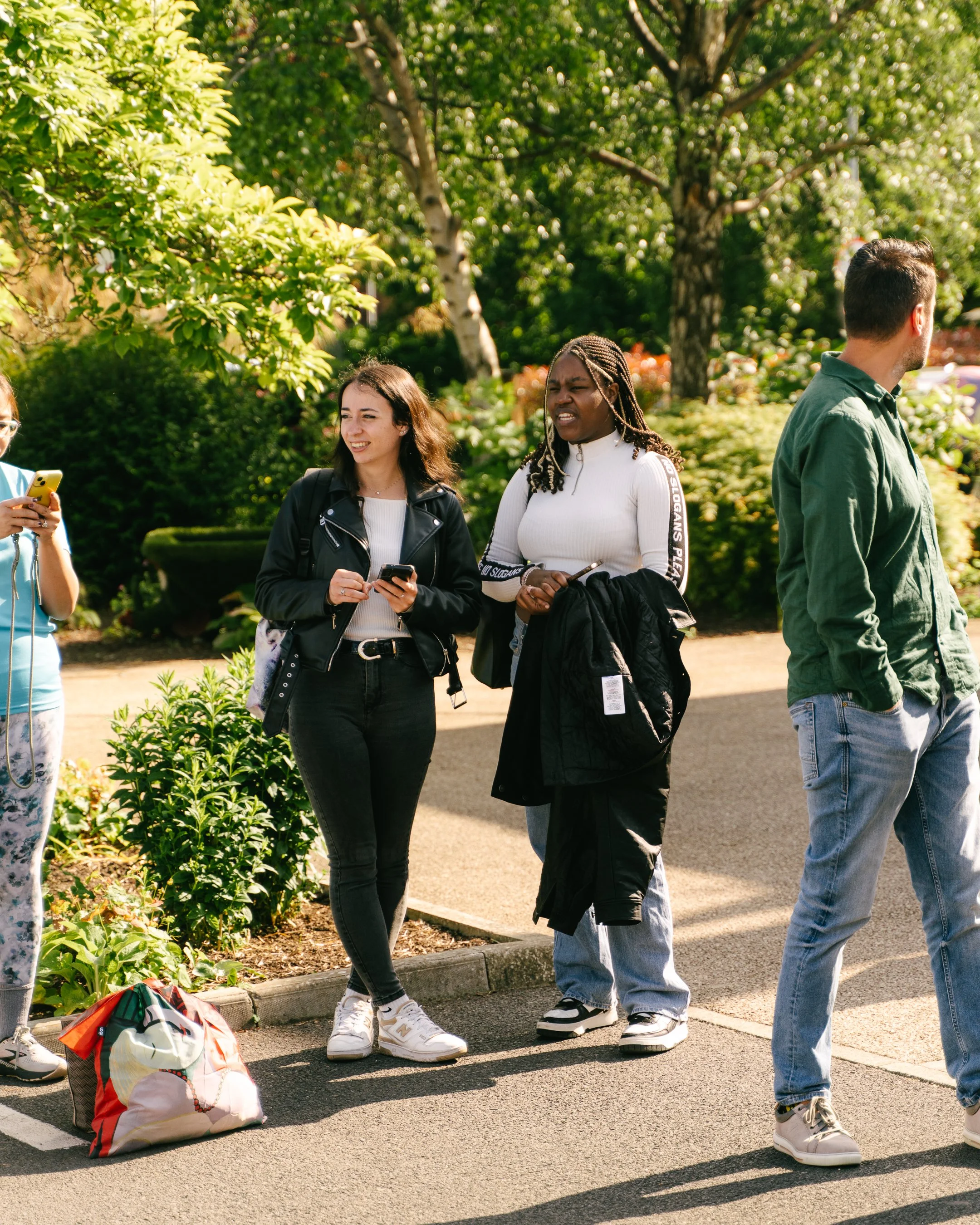 People standing on the sidewalk in a park, with a woman looking at her phone, another woman holding a black jacket, and a man looking away, surrounded by green trees and bushes.