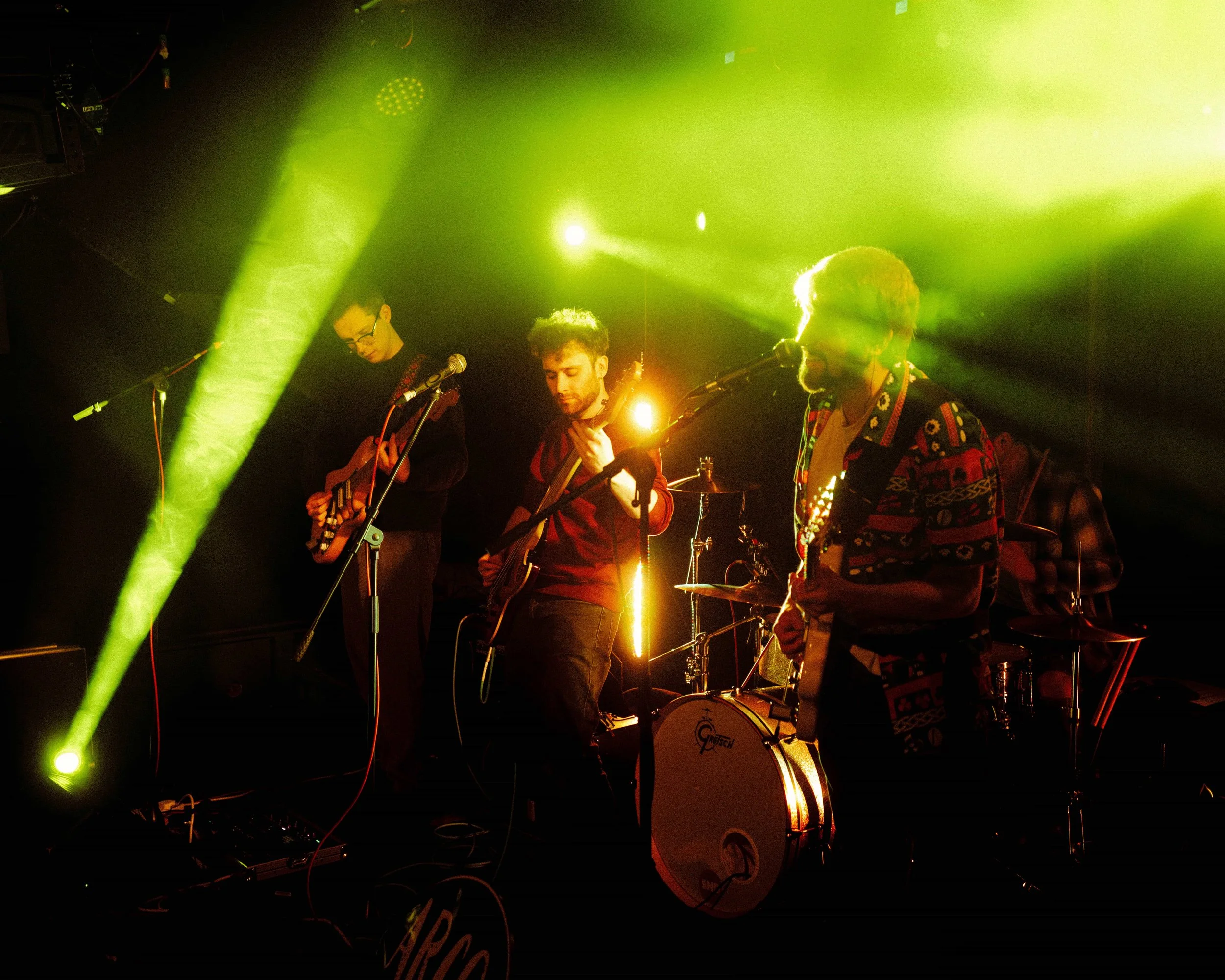 Band performing on stage with green and yellow lights shining on them, playing guitars and drums.