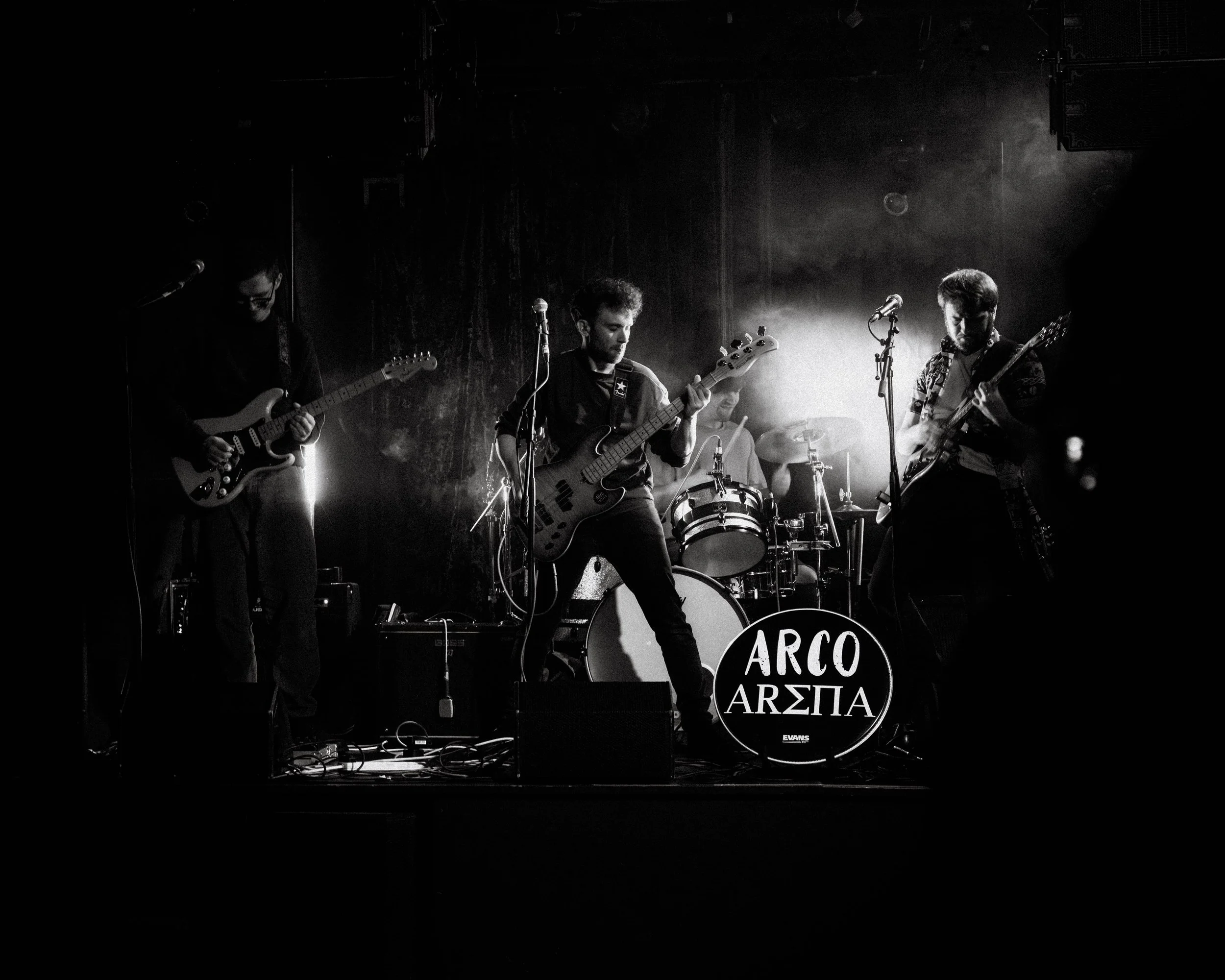 A four-piece band performing on stage with guitars, bass, and drums. The drum set has a logo with the words 'ARCO ARΣA'. The scene is dark with spotlights illuminating the musicians.