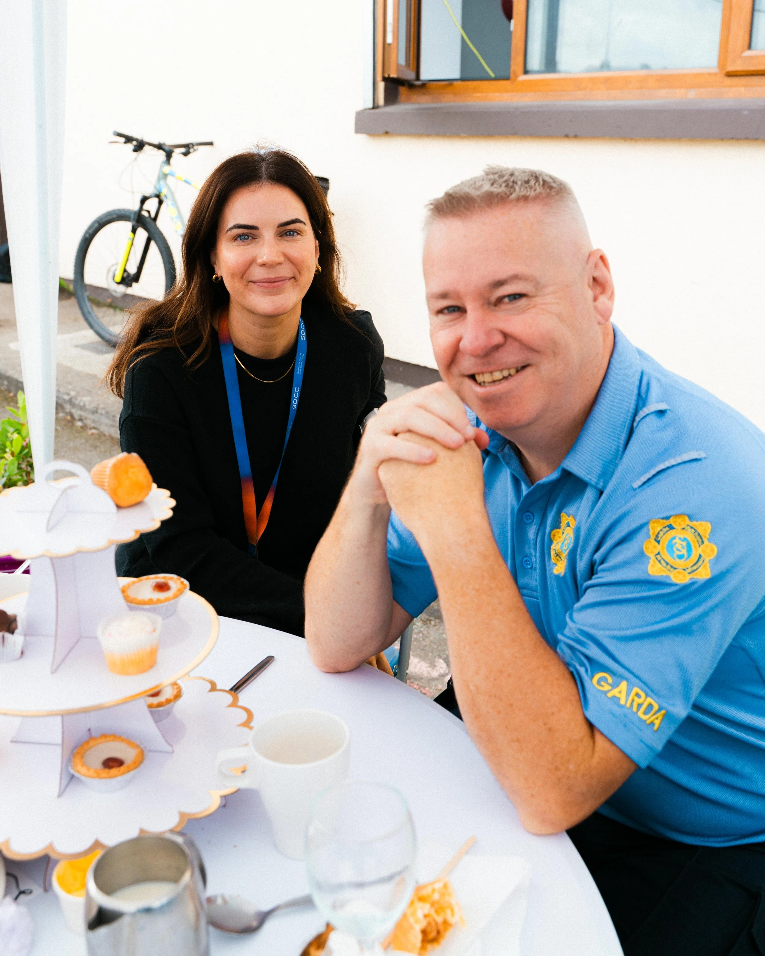 Two smiling people sitting at a table with a tiered stand of cupcakes, outdoors in front of a white wall with a window and a bicycle in the background.