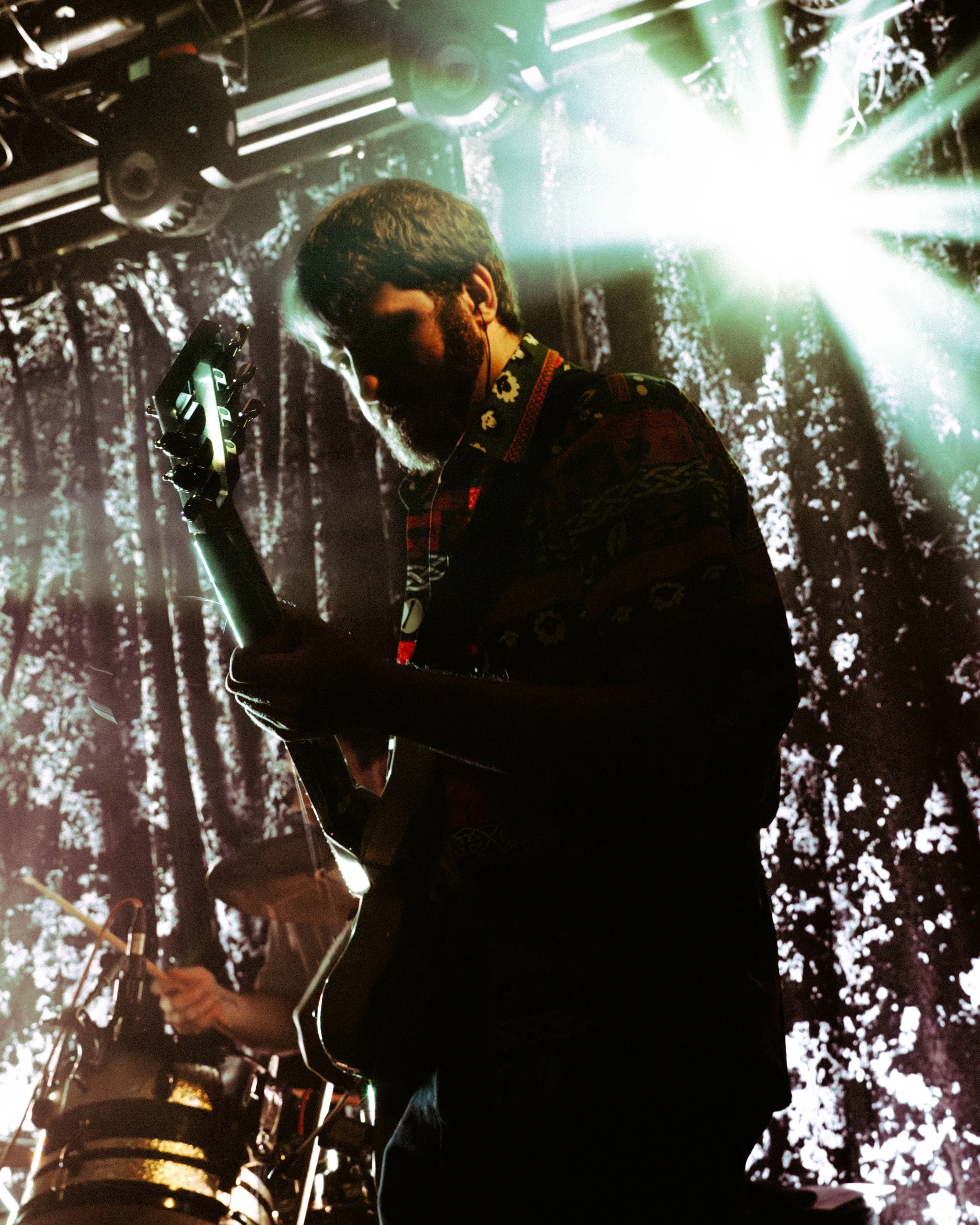 A musician playing an electric guitar on stage with a backdrop of trees and bright stage lights.