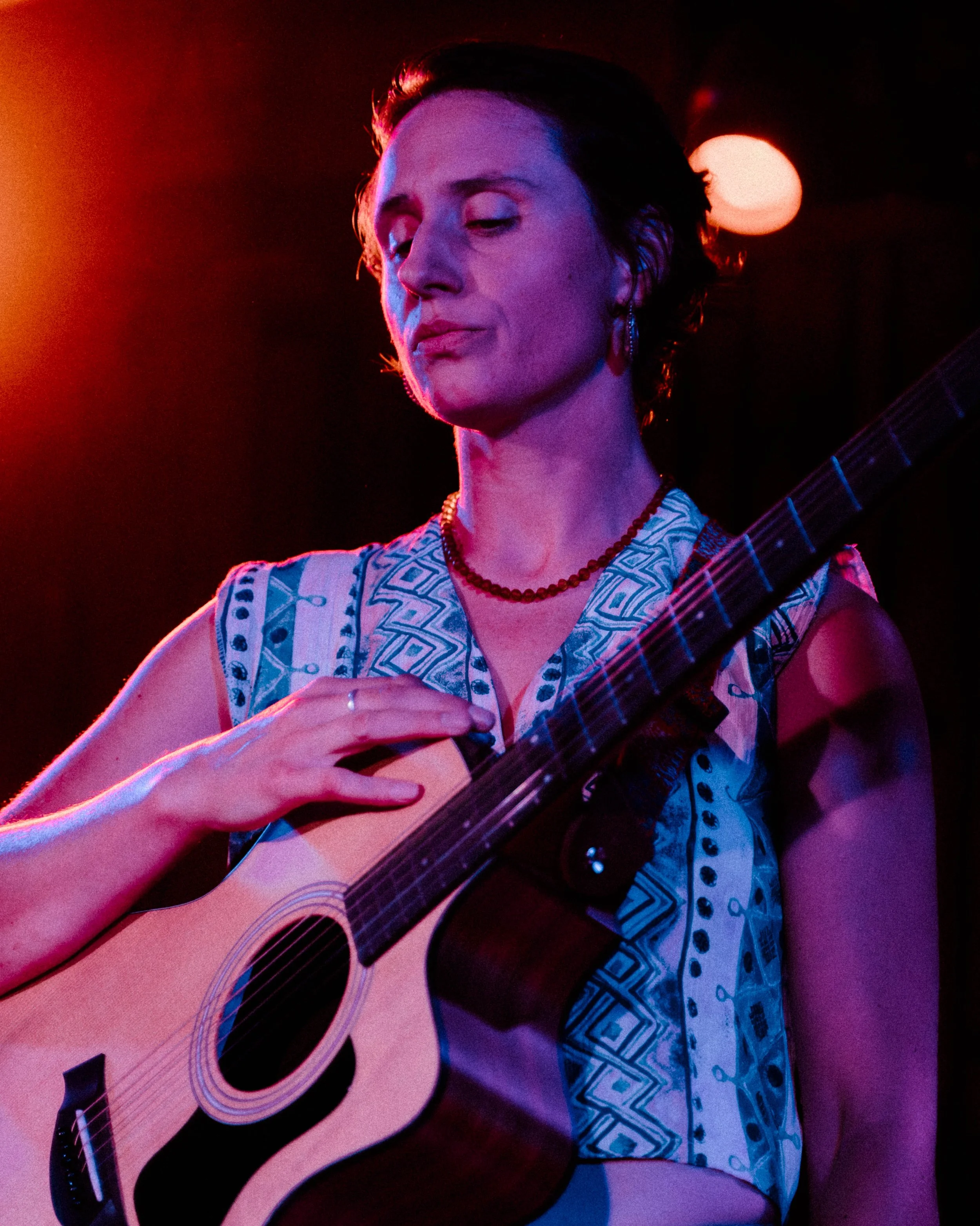 A woman with short dark hair playing an acoustic guitar, wearing patterned clothing, earrings, a necklace, and has closed eyes in a dimly lit setting with colorful lighting.