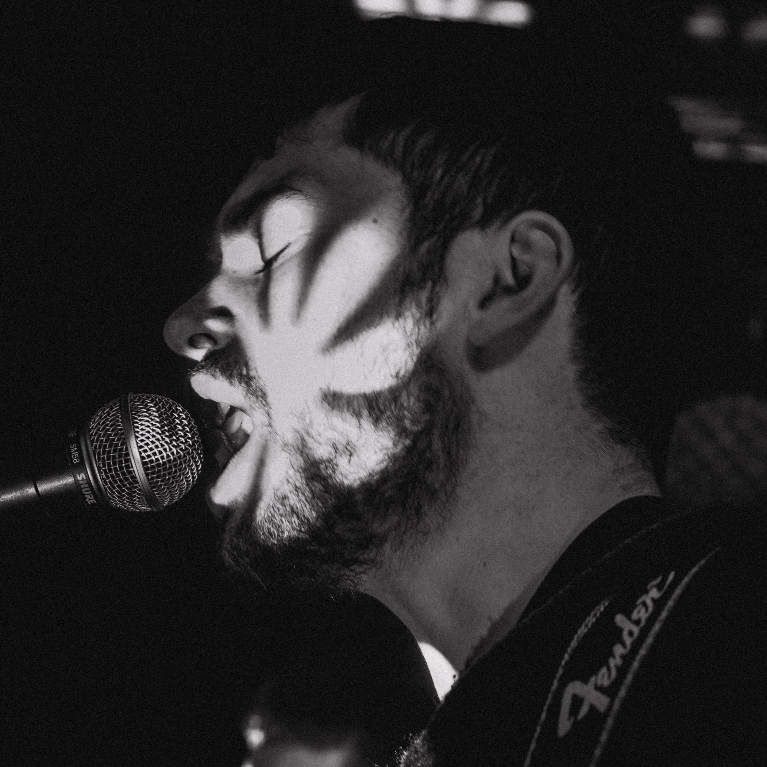 Black and white photo of a man singing into a microphone, with shadows and dramatic lighting on his face.