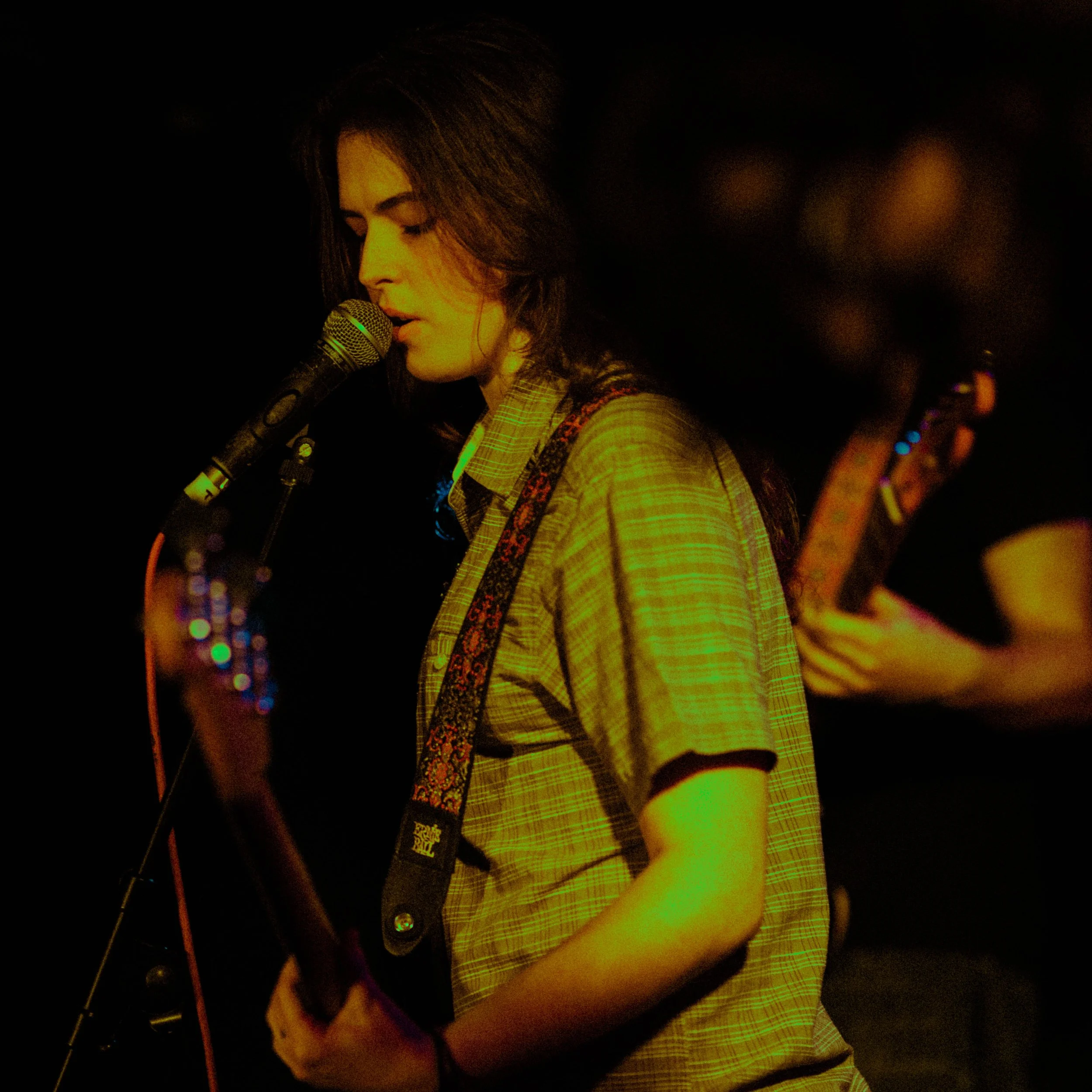 A young woman with brown hair, wearing a beige plaid shirt, singing into a microphone while playing an electric guitar on stage at night.