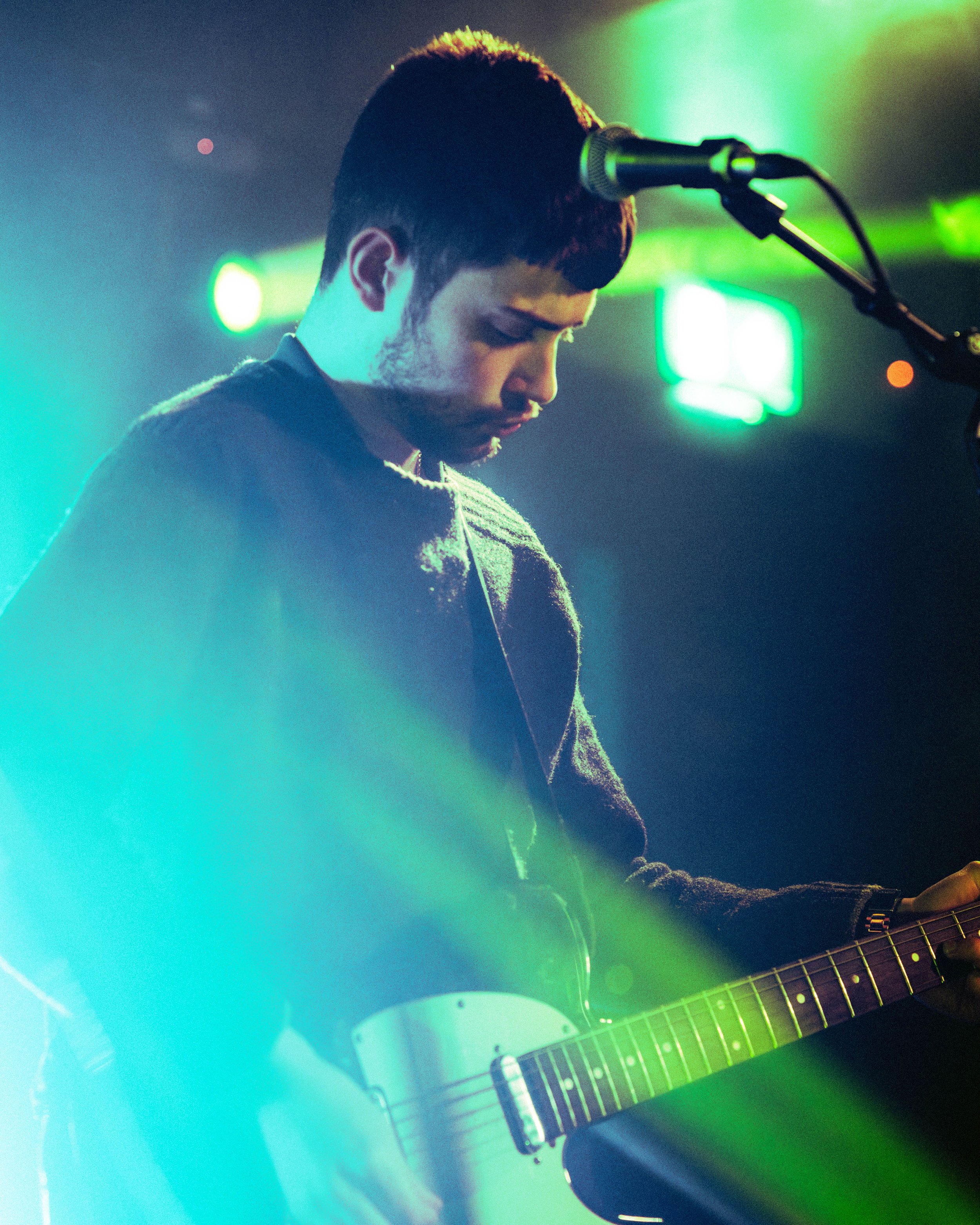A young man playing an electric guitar with colorful stage lighting and a microphone in front of him.