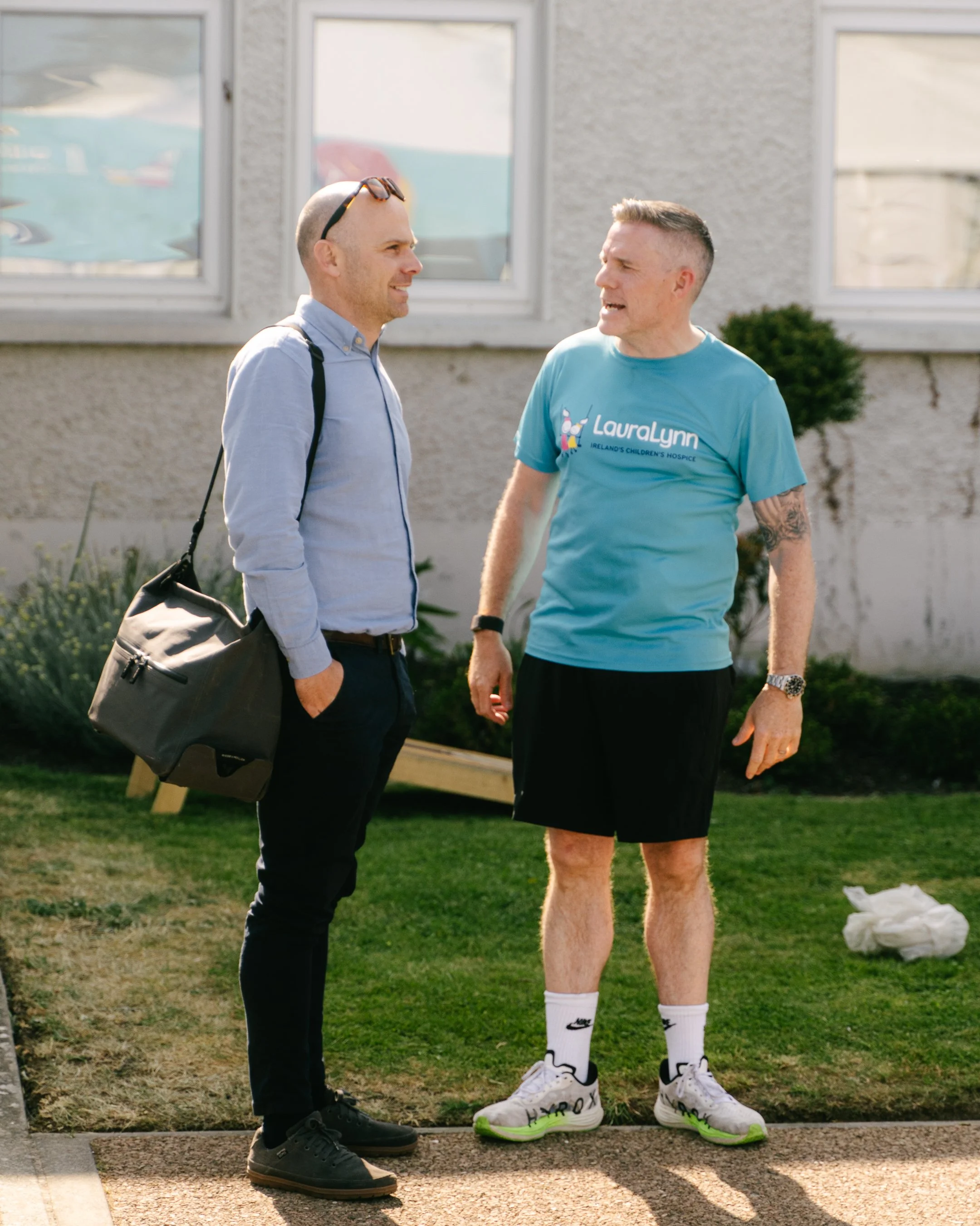Two men having a conversation outdoors in front of a building. One man is wearing a light blue shirt and black pants with a shoulder bag, and the other is dressed in a light blue T-shirt, black shorts, white socks, and sneakers. The man in casual wea