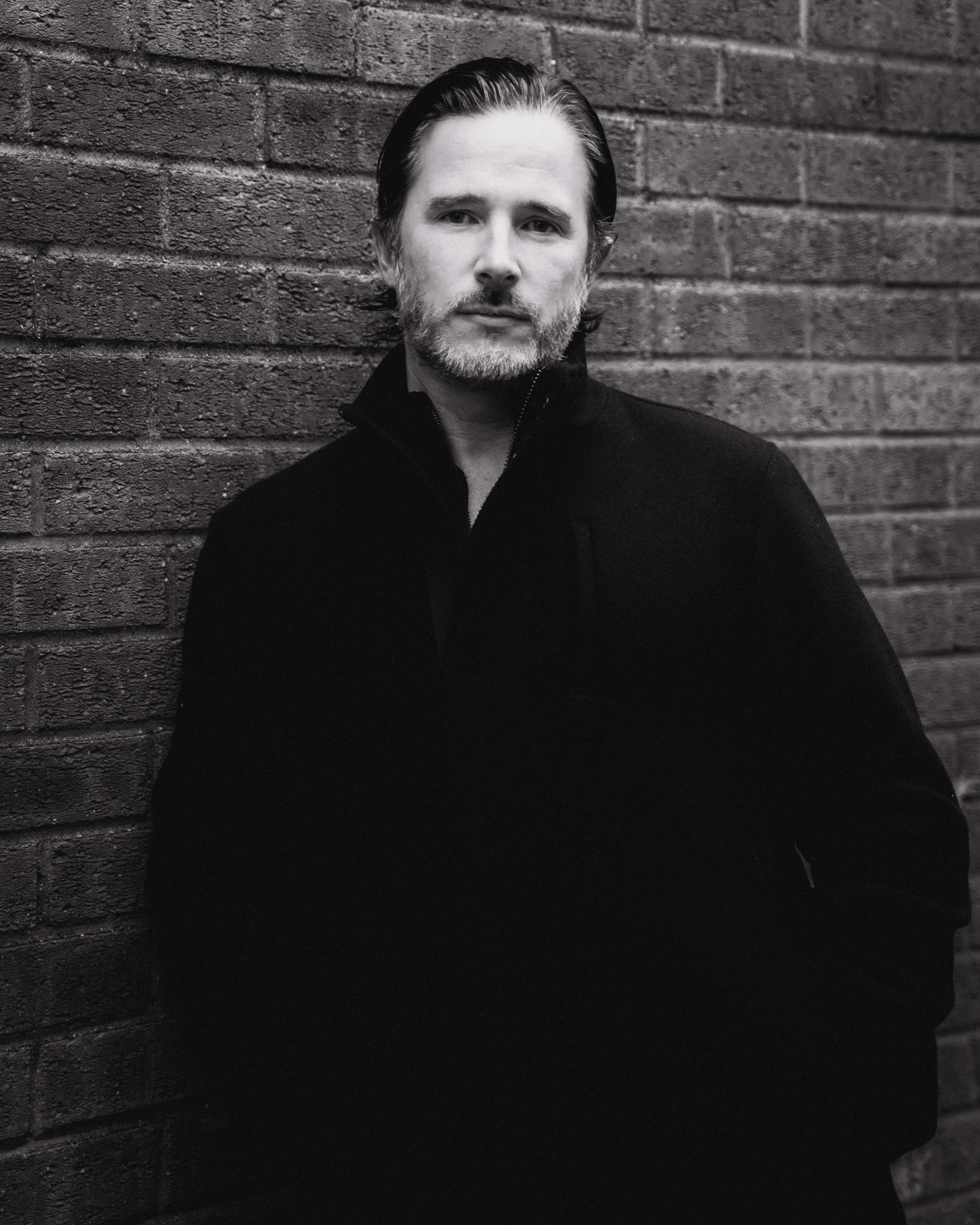 Black and white portrait of a man with a beard, slicked-back hair, wearing a dark jacket, standing against a brick wall.