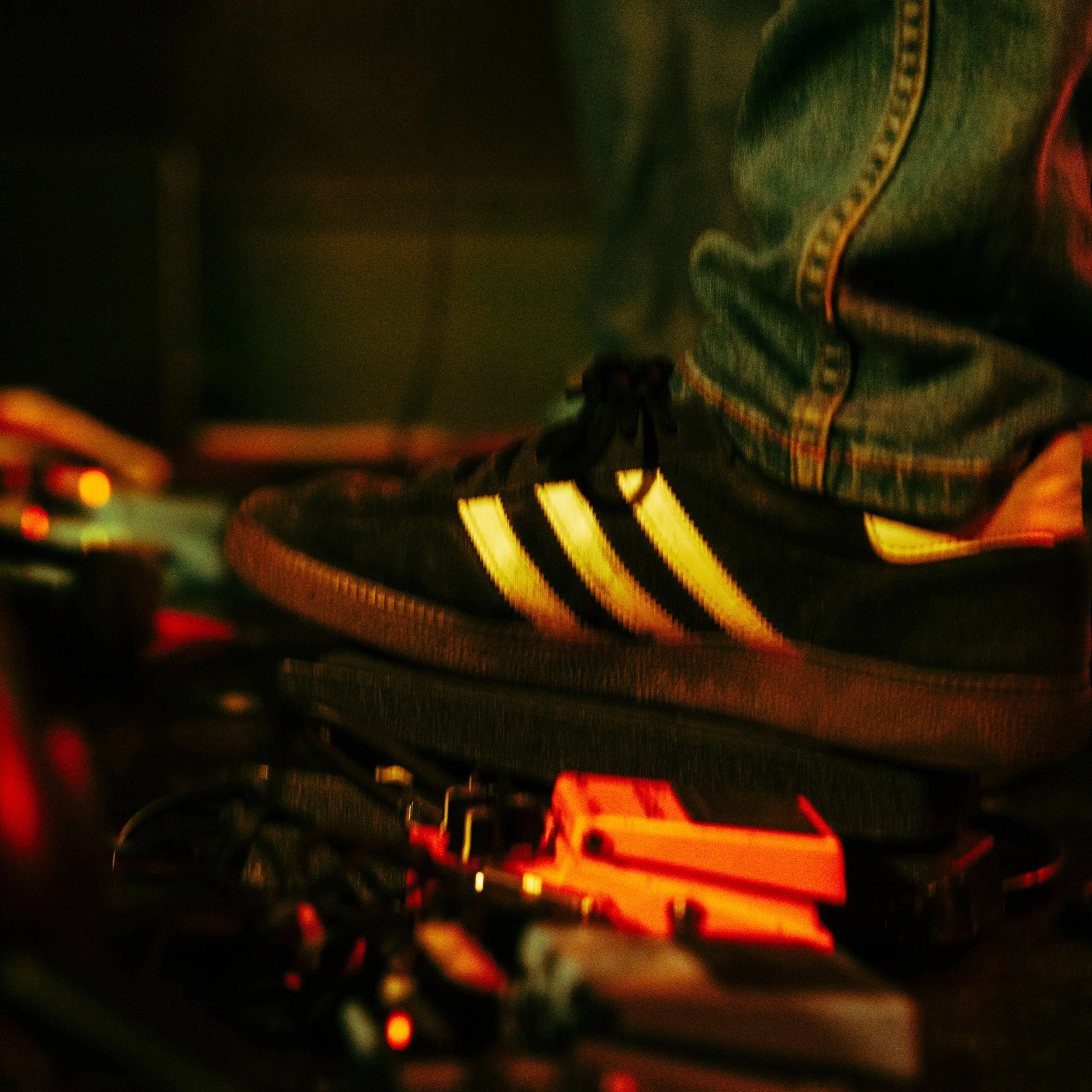 Close-up of a black sneaker with three white stripes and a white sole, worn with jeans, on a pedal board with guitar effects pedals underneath, in a dimly lit environment.