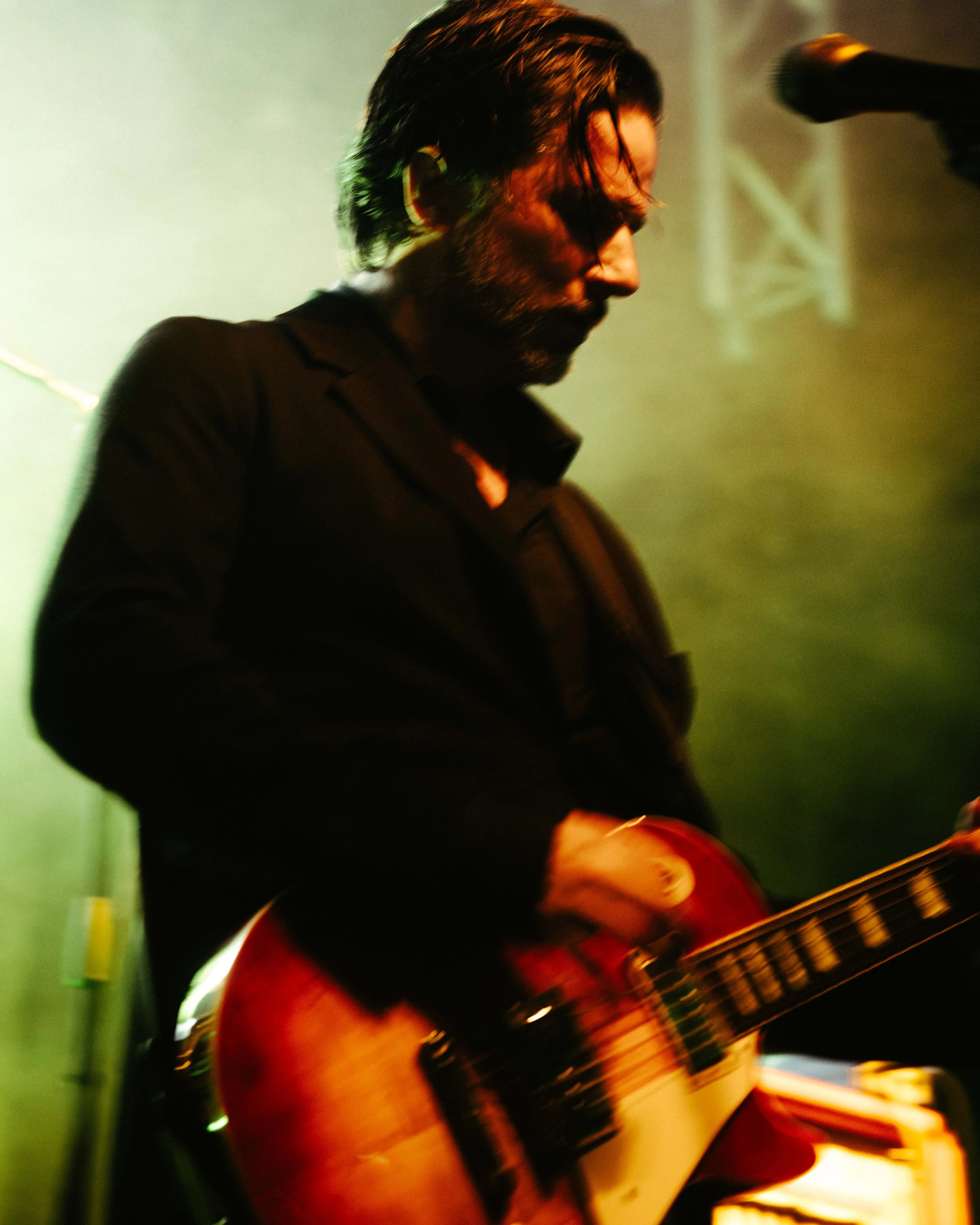 A man with dark hair and beard playing an electric guitar on stage, wearing dark clothing, with stage lighting creating a moody atmosphere.