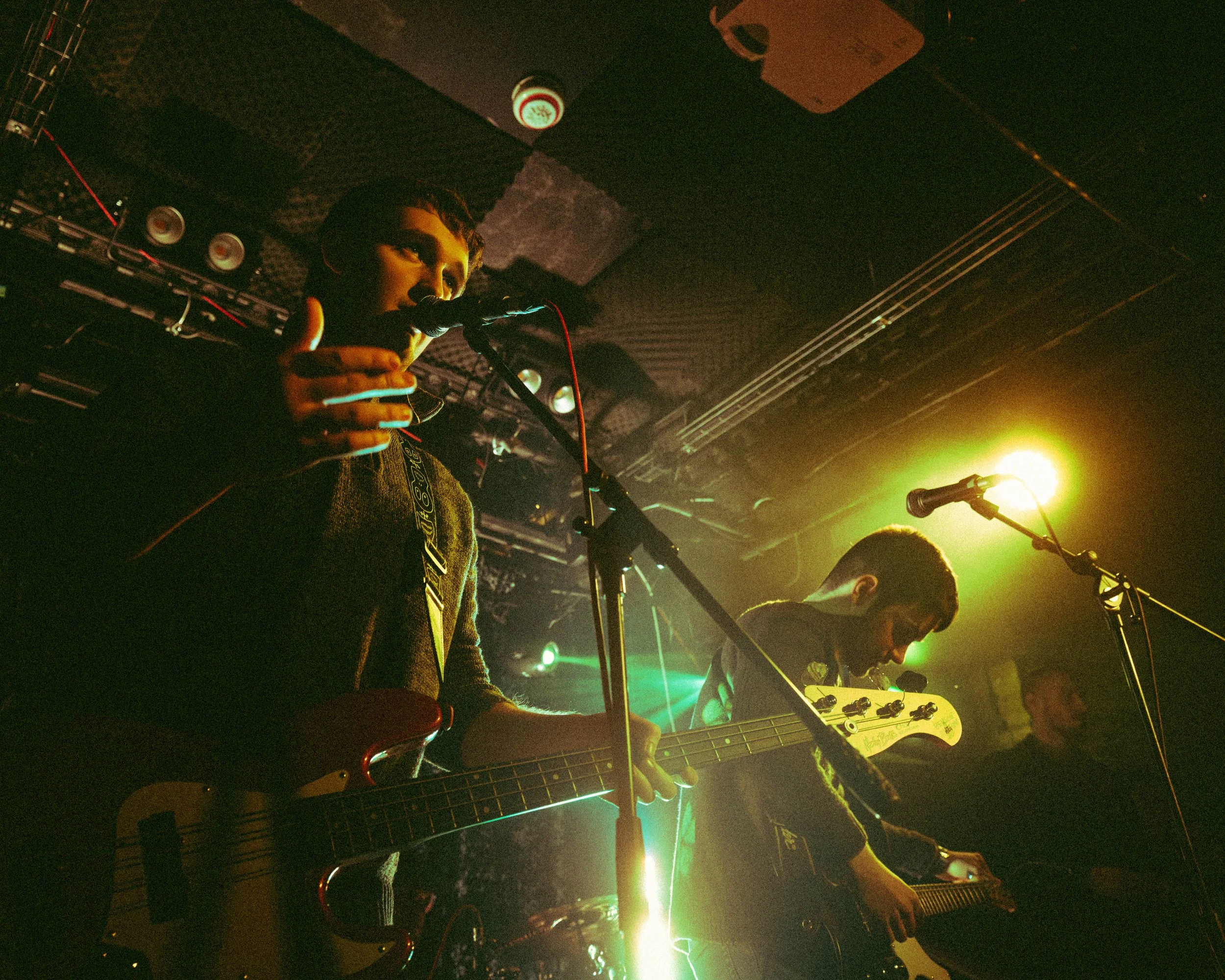 Two musicians perform on stage with guitars, under colorful concert lighting, in a dark venue.