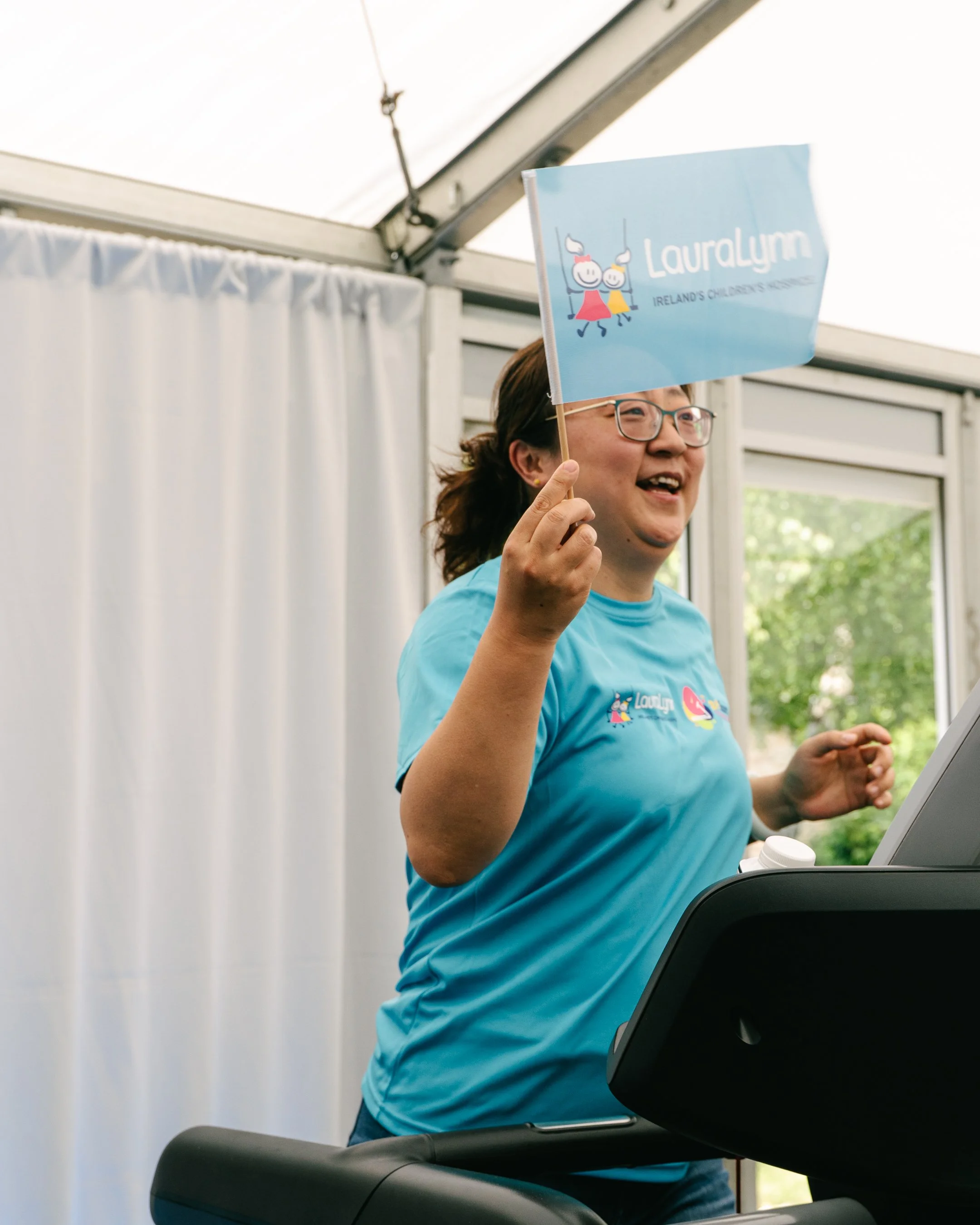 A woman with glasses and a blue shirt holding a blue flag with cartoon characters and the text 'LauraLynn Ireland's Children's Hospice' in a bright indoor setting.
