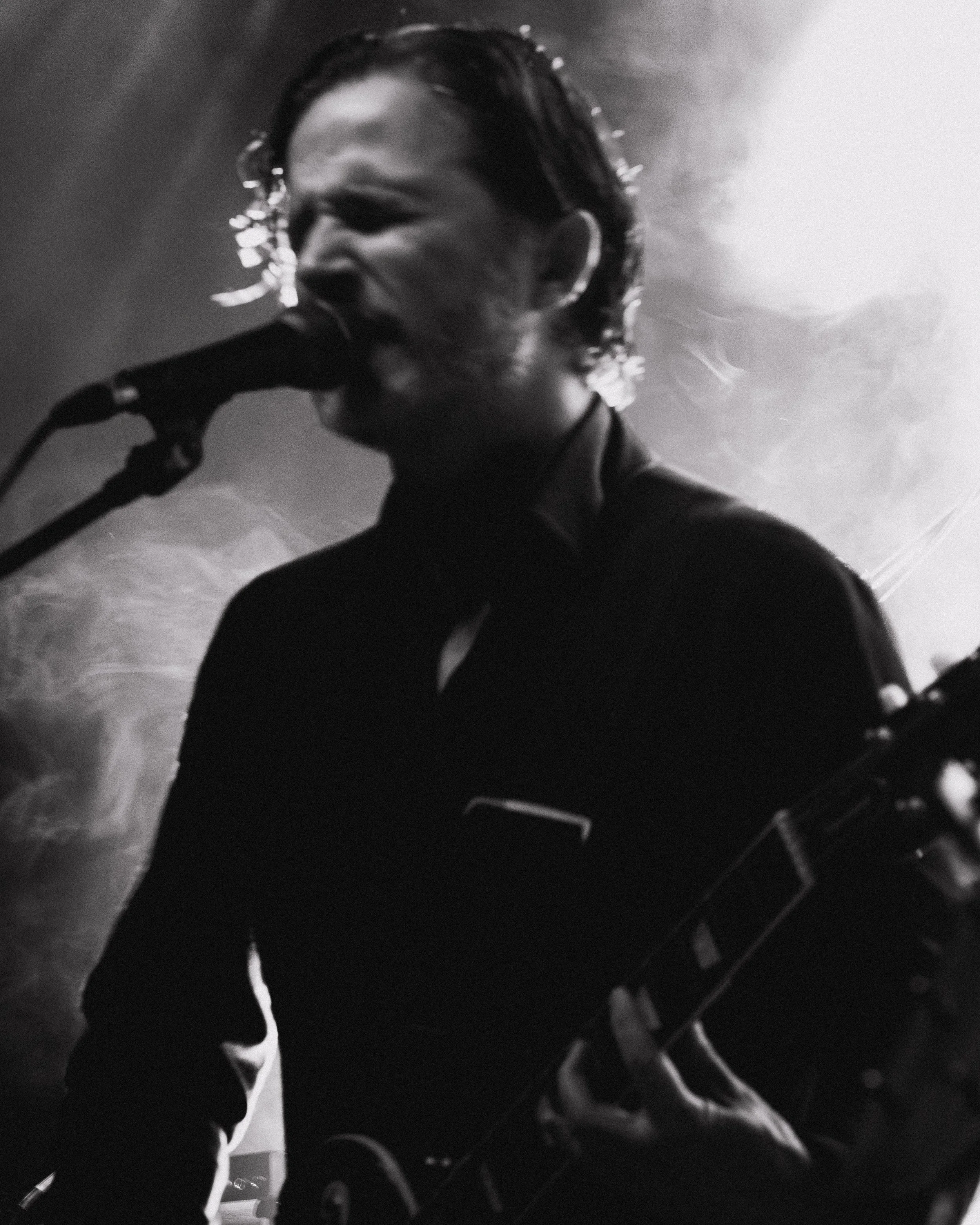Black and white image of a male musician singing into a microphone and playing an electric guitar with smoke and stage lighting in the background.