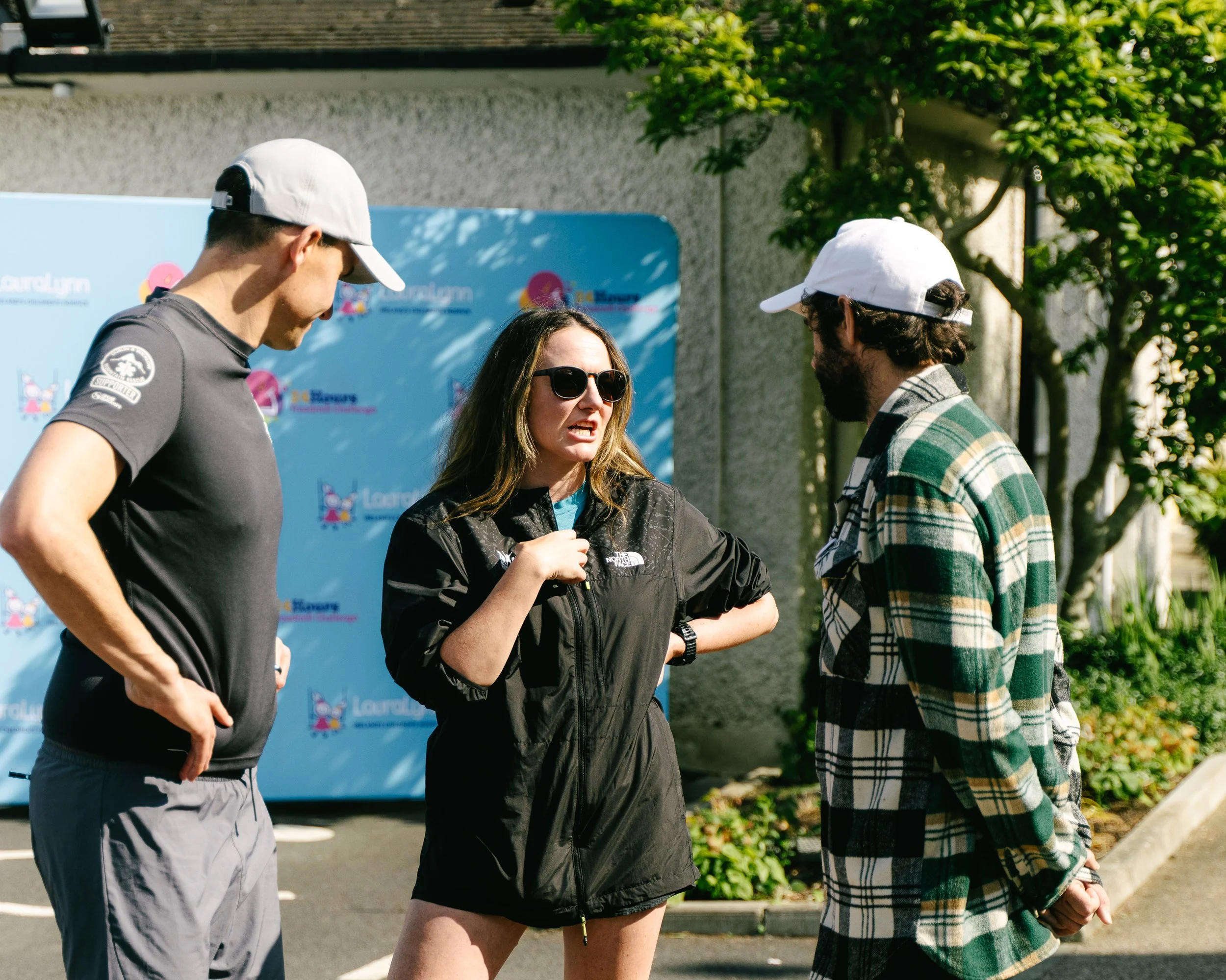 Four people standing outdoors near a blue backdrop with logos, engaged in conversation. One woman is wearing sunglasses and a black jacket, speaking to two men in caps and casual clothing, one of whom is wearing a plaid jacket. Green trees and a gray