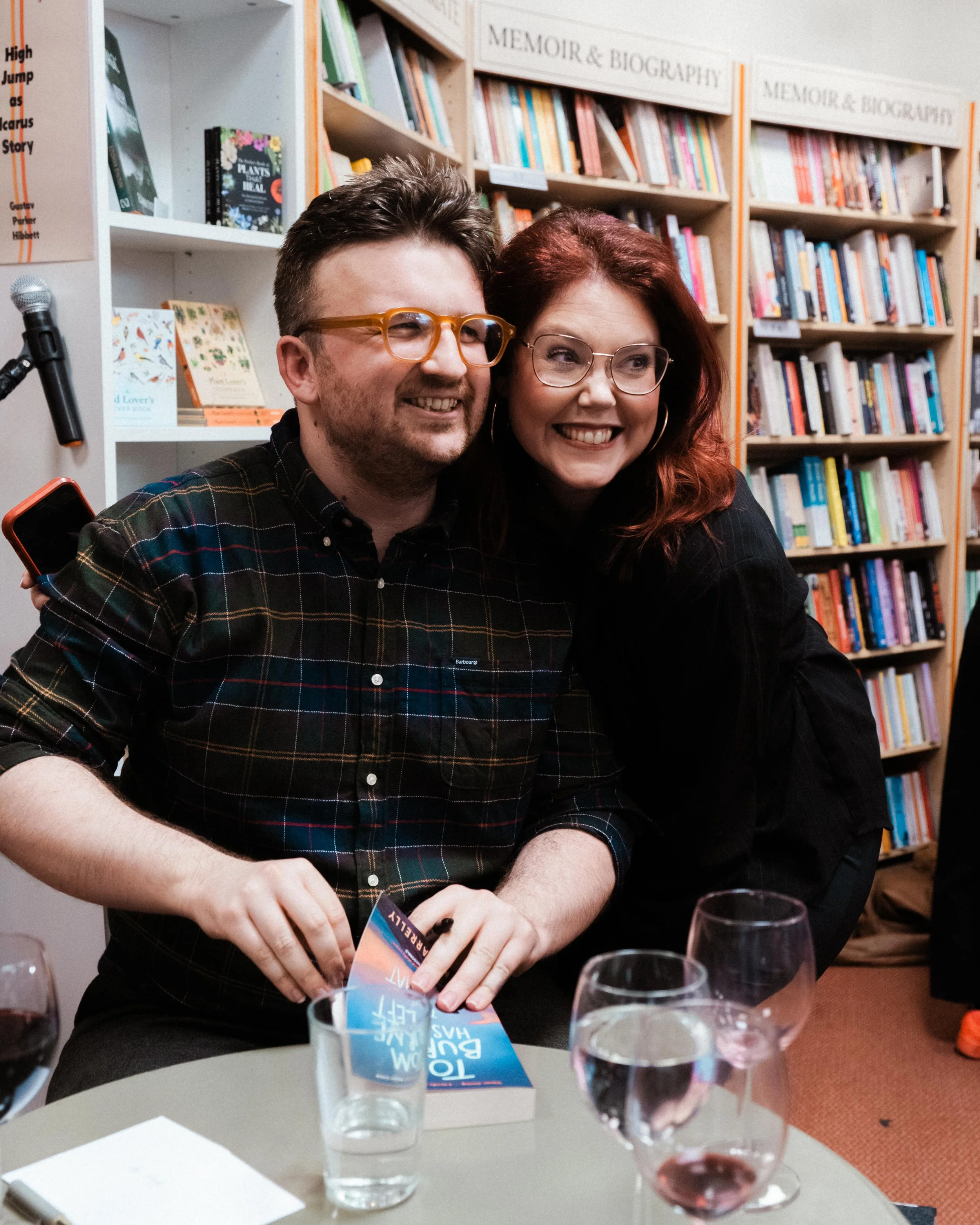 Two smiling people, a man with glasses and a woman, are at a book event or signing. They are standing in front of bookshelves labeled 'Memoir & Biography' and appear to be posing for a photo. The man is holding a book and is seated at a table with se