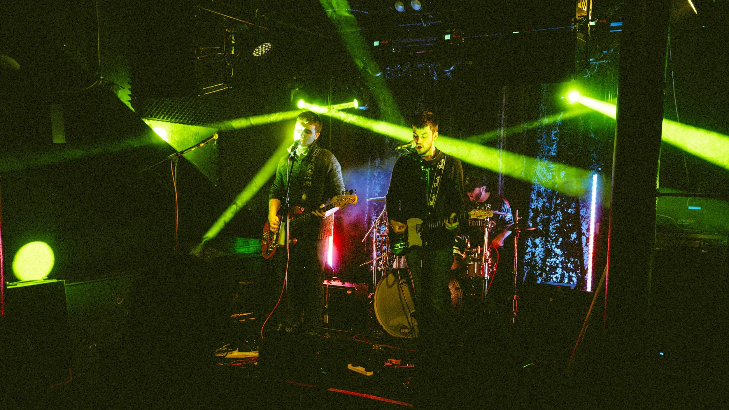 A four-member band performing on stage with green stage lights and a dark background. Two guitarists are at front, singing into microphones, with a drummer in the background.