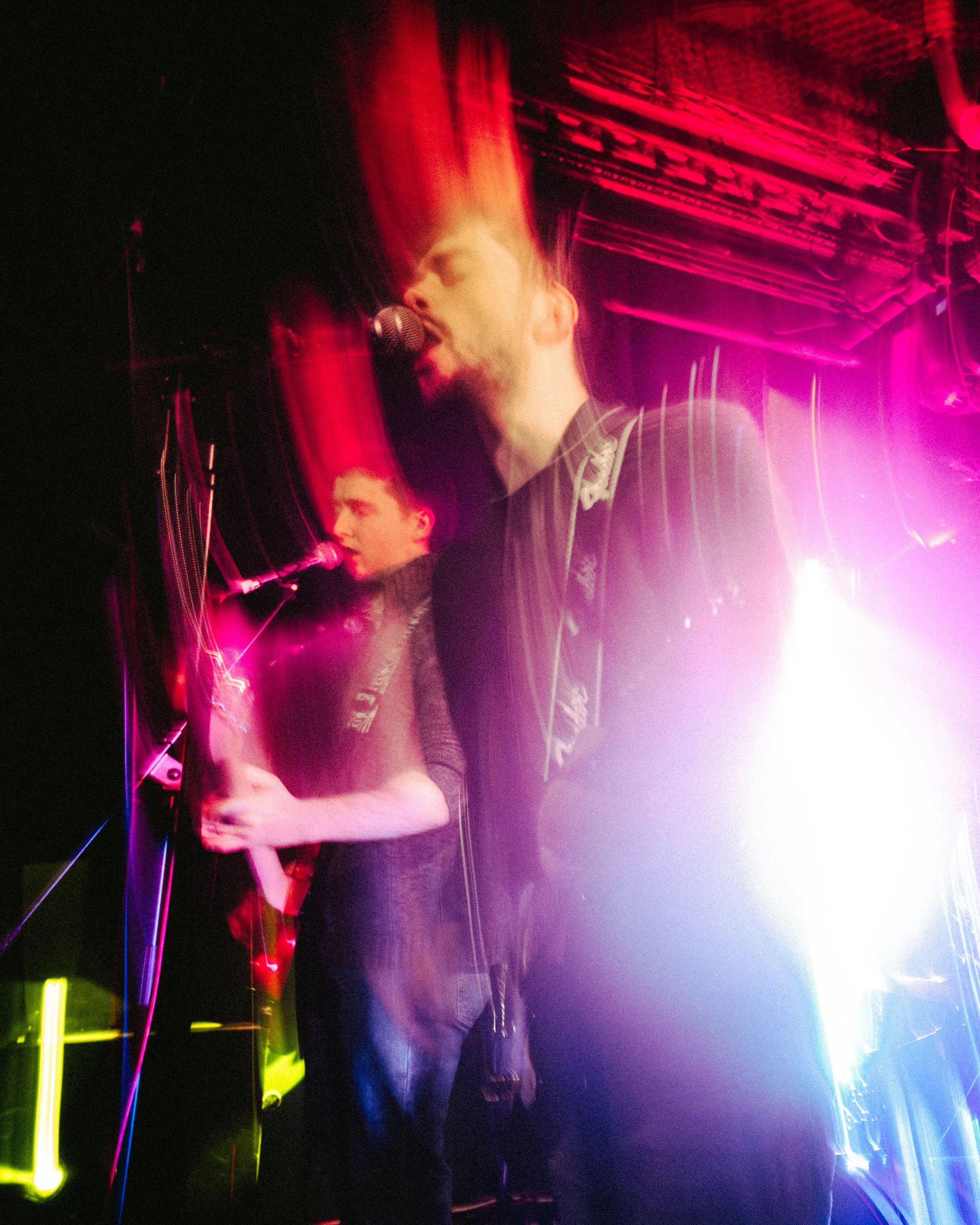 Two musicians performing on stage with neon lighting, one singing into a microphone with motion blur and light trails, the other playing an instrument in the background.