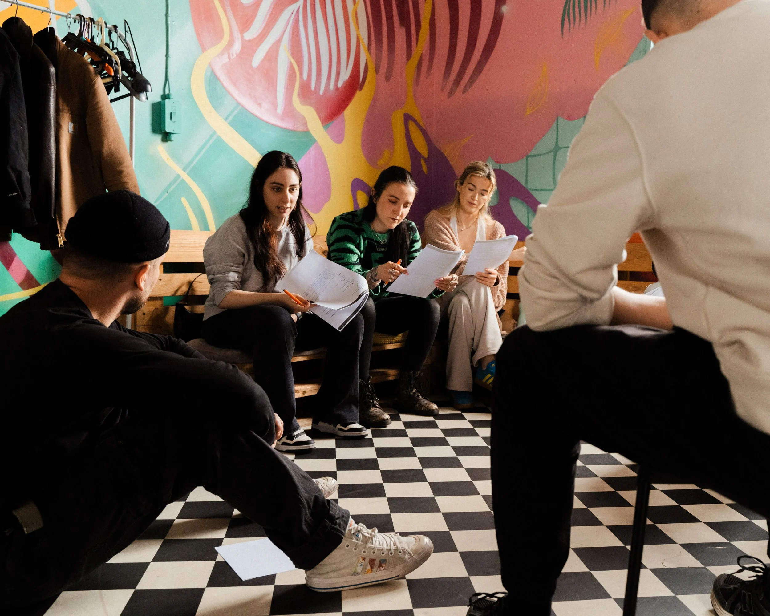 People sitting at a table with documents and pens, in front of a colorful, abstract wall mural, with a person standing and talking, and a clothing rack with jackets on the side.