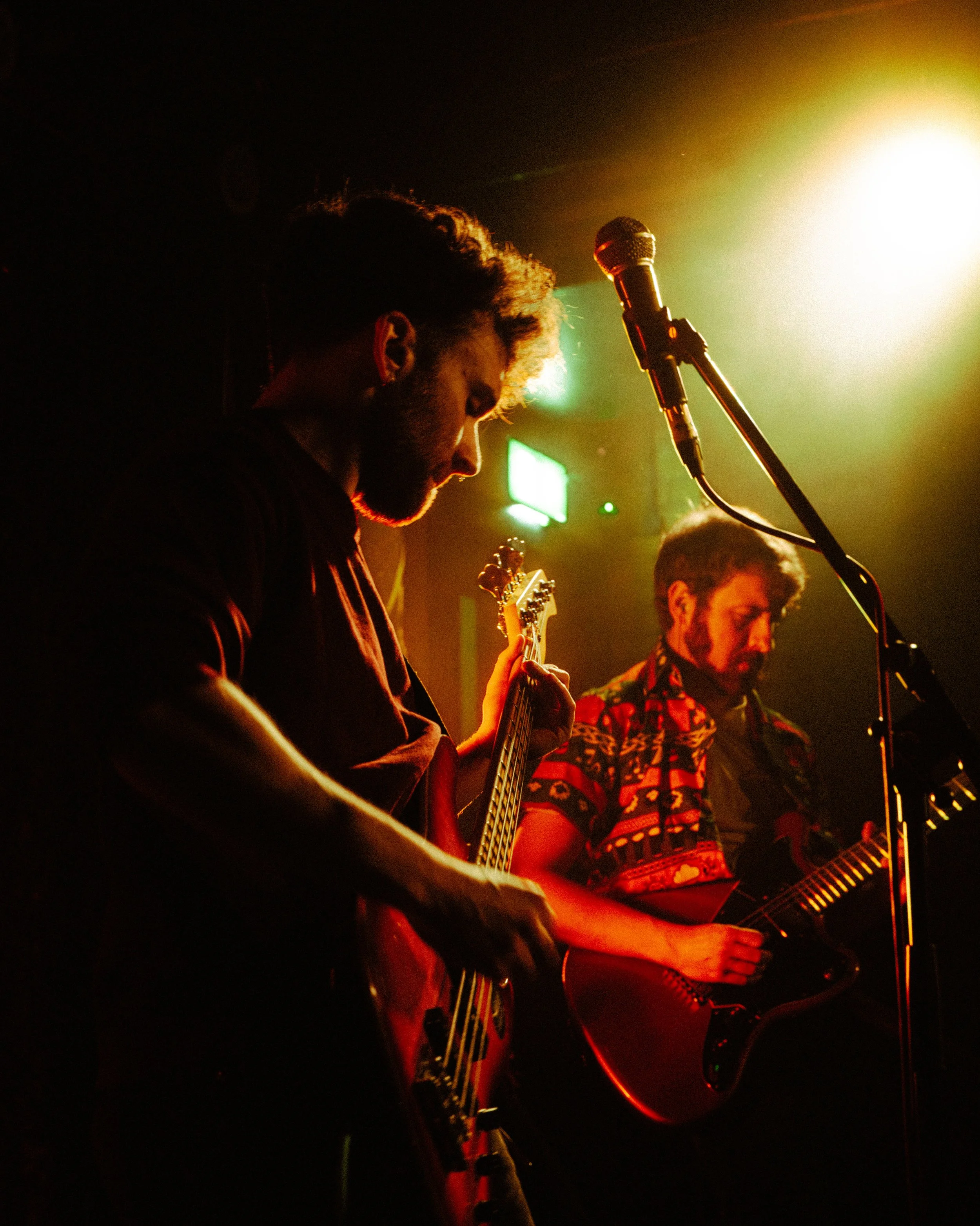 Two male musicians performing on stage with guitars under warm stage lighting.