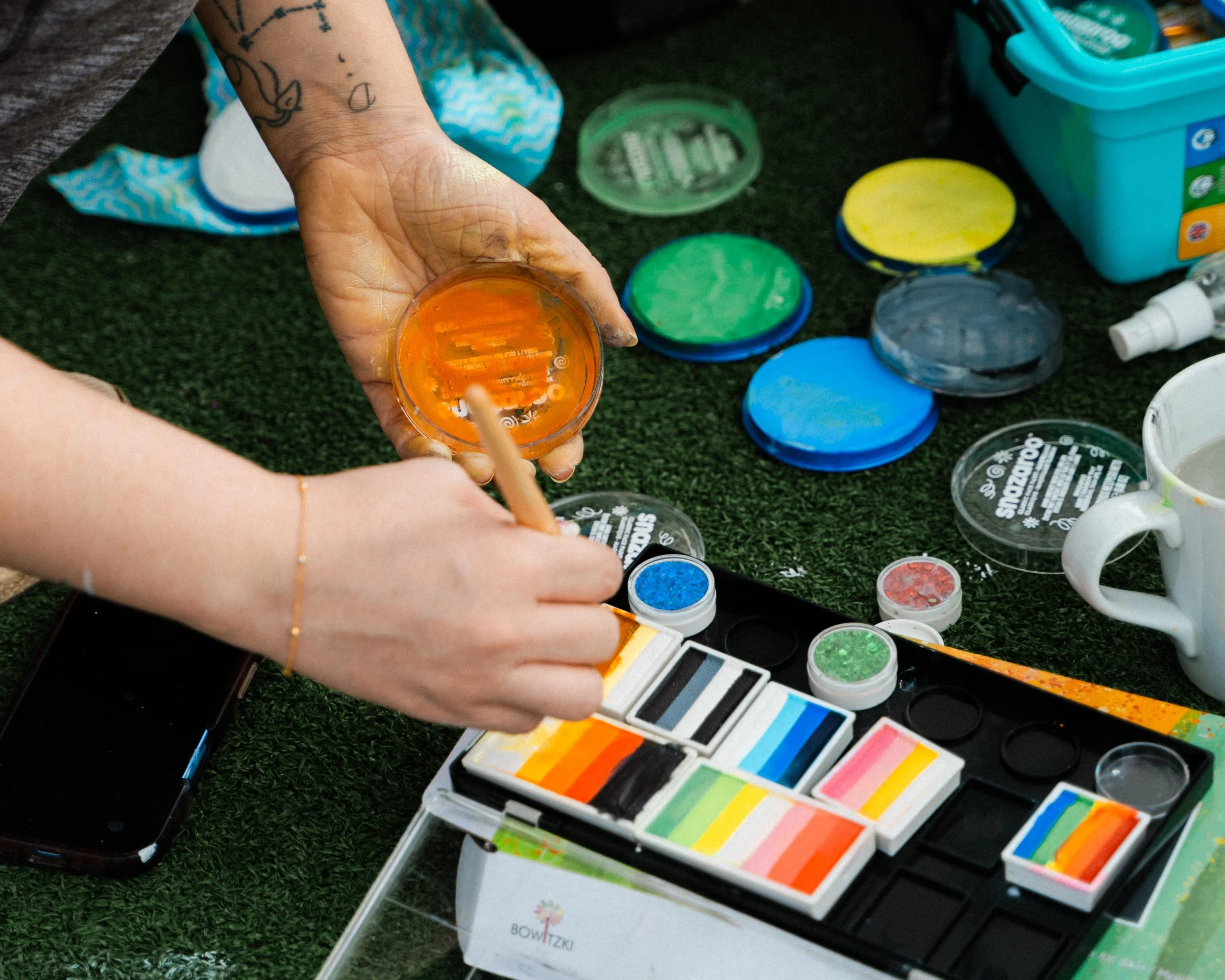 Person handling a small container of orange slime with a wooden stick, surrounded by various colorful slime and arts and crafts supplies on a black surface.